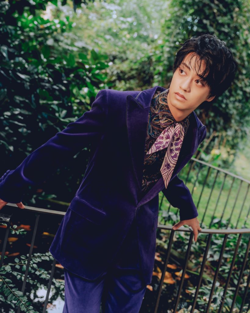 A person wearing a purple velvet jacket with a patterned shirt leans on a railing in a lush garden setting, looking thoughtfully off to the side.