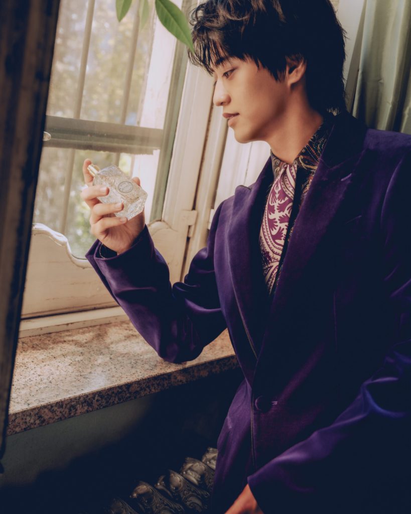 A man dressed in a purple velvet blazer and patterned shirt holds a perfume bottle while gazing out a window, surrounded by soft natural light and greenery.