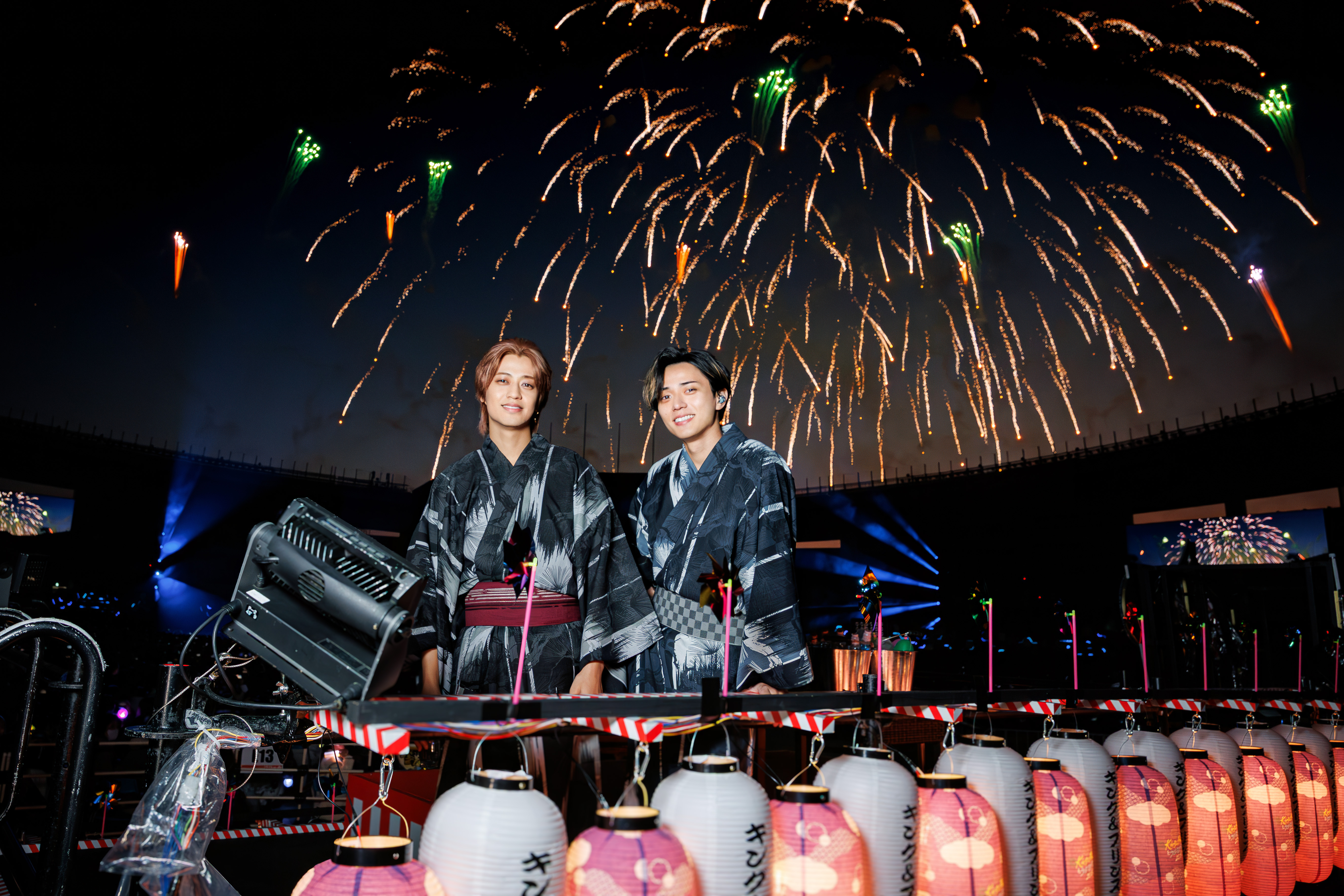 Two performers dressed in traditional yukata stand in front of a backdrop of colorful fireworks at a night event, with decorative lanterns in the foreground.