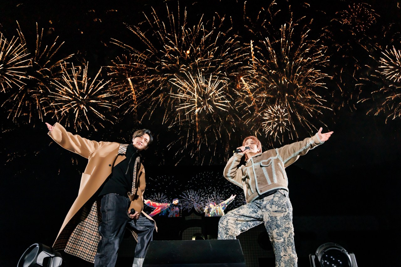 Two performers on stage smiling and singing, with a backdrop of vibrant fireworks lighting up the night sky.