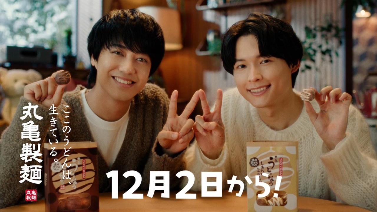 Two young men smiling and holding a Marugame Udon product in a cozy, indoor setting with a winter theme.
