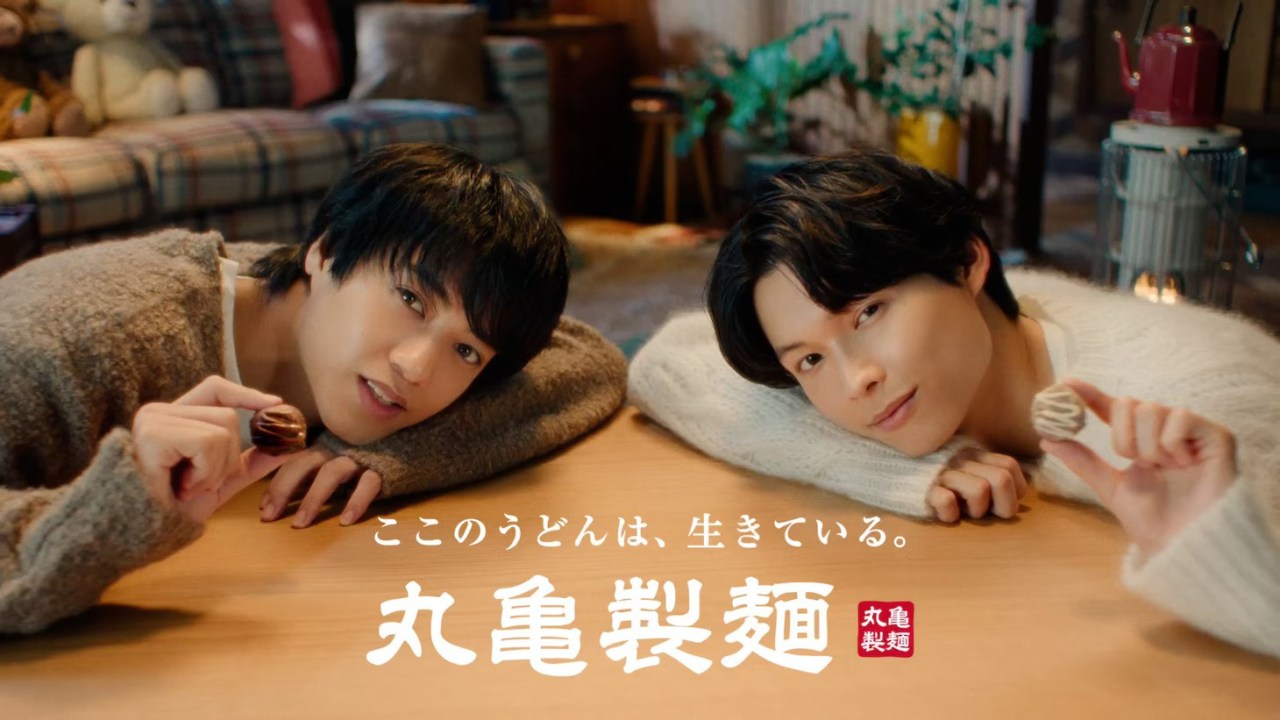 Two young men sitting at a table in a cozy setting, with one holding a chocolate treat, both looking at the camera with friendly expressions. The background features a warmly decorated room with plush toys and soft furnishings.