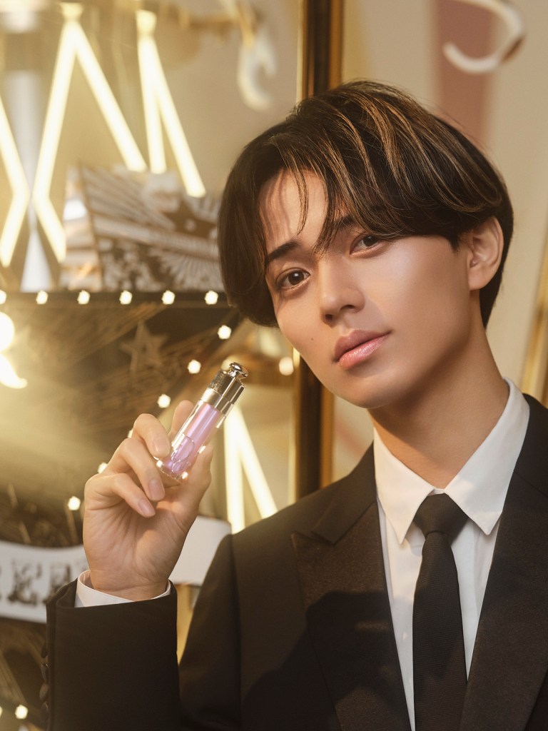 A young man in a black suit holds a pink lipstick product, posing against a backdrop featuring sparkling lights and decorative elements.