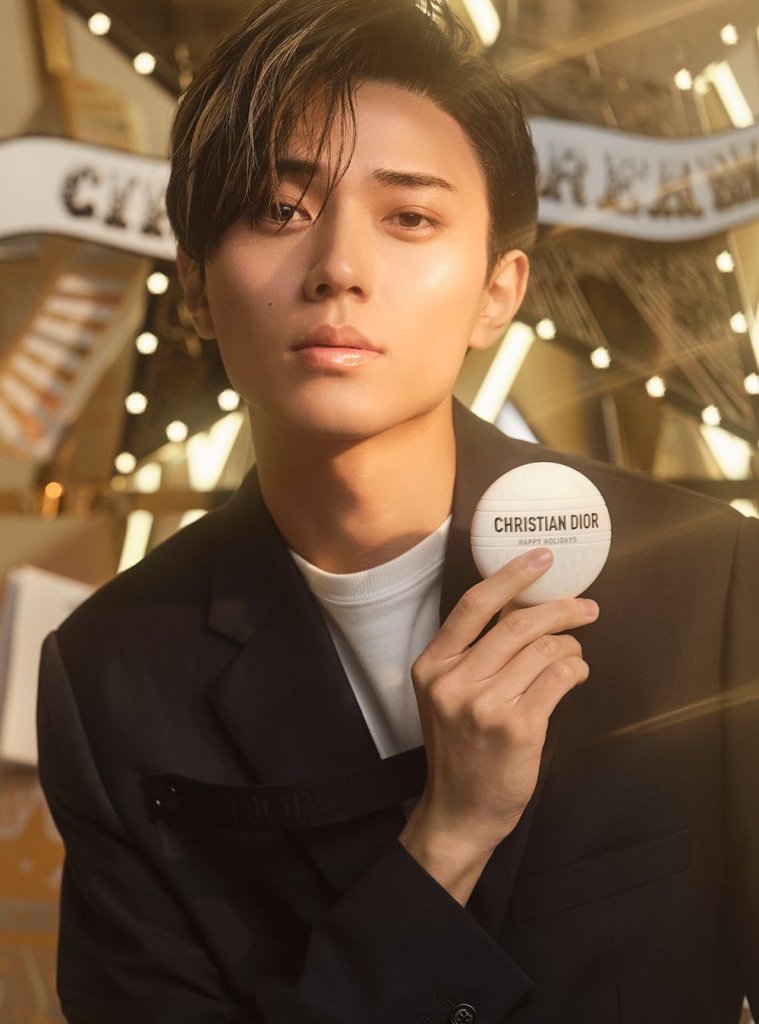 A close-up of a stylish young man holding a Christian Dior product, with a warm, illuminated background reflecting a festive atmosphere.