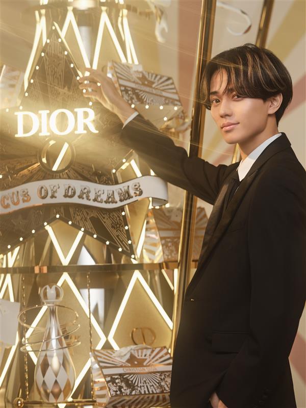 Ren Nagase posing elegantly beside a Dior display featuring a star that reads 'CIRCUS OF DREAMS'. He is dressed in a black suit with a white shirt, showcasing a stylish demeanor against a glamorous backdrop.