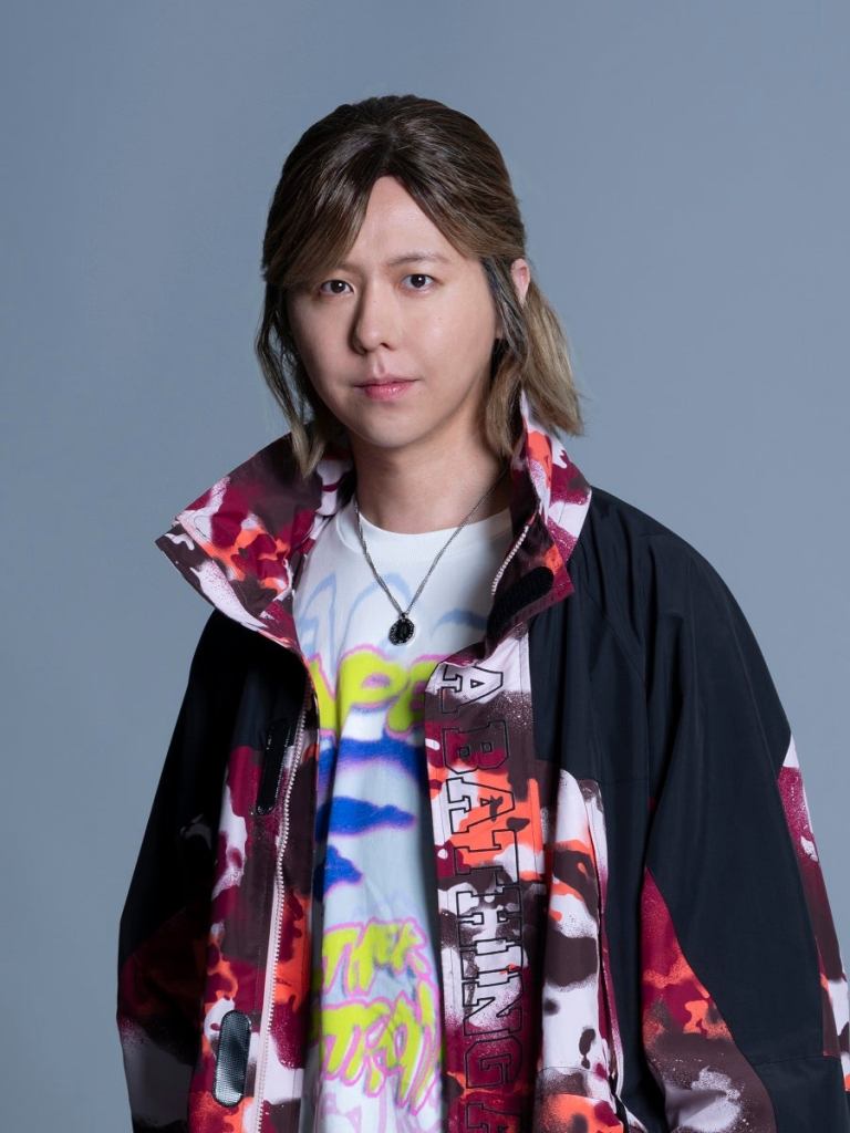 An individual wearing a colorful jacket and t-shirt, posing in front of a plain background.