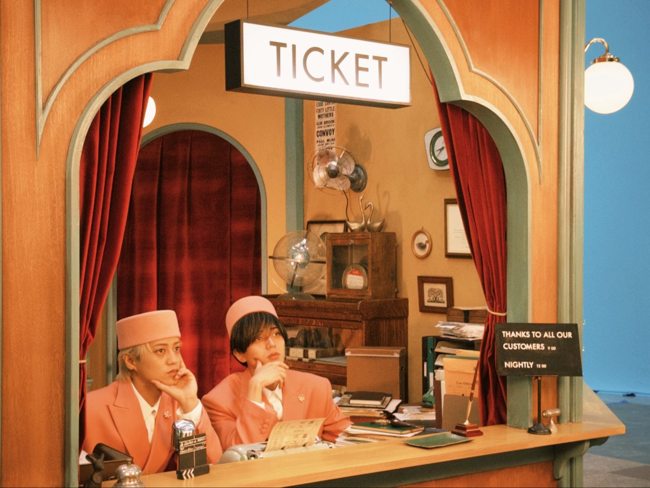 Two individuals dressed in pink uniforms sit at a vintage ticket booth, with a sign reading 'TICKET' above them, set against a retro backdrop featuring decorative elements.
