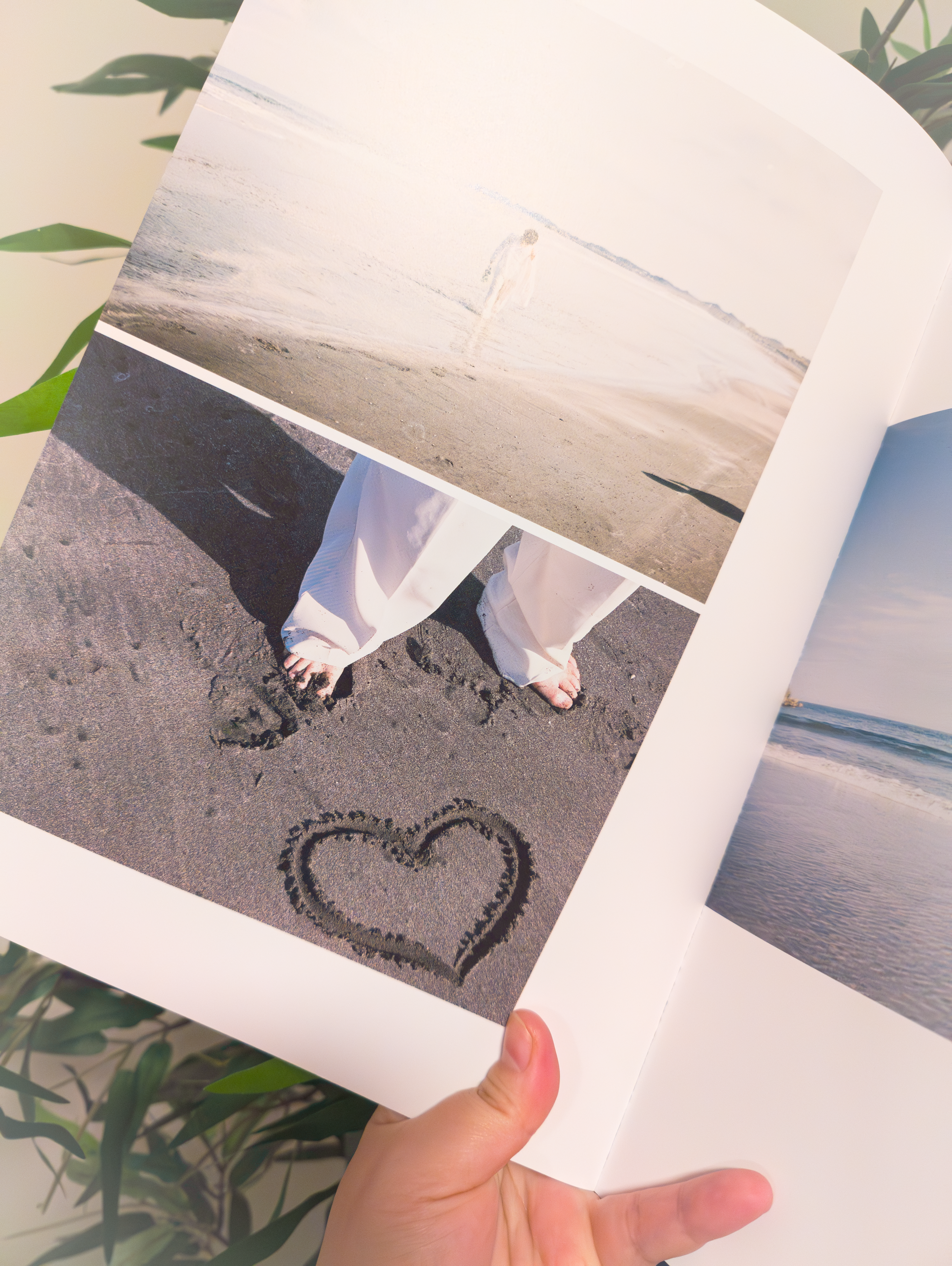 A hand holds an open photobook showing three images on a page: a figure walking on a beach, close-up of sandy feet beside a heart drawn in the sand, and a view of the sea. In the background, green leaves are partially visible.