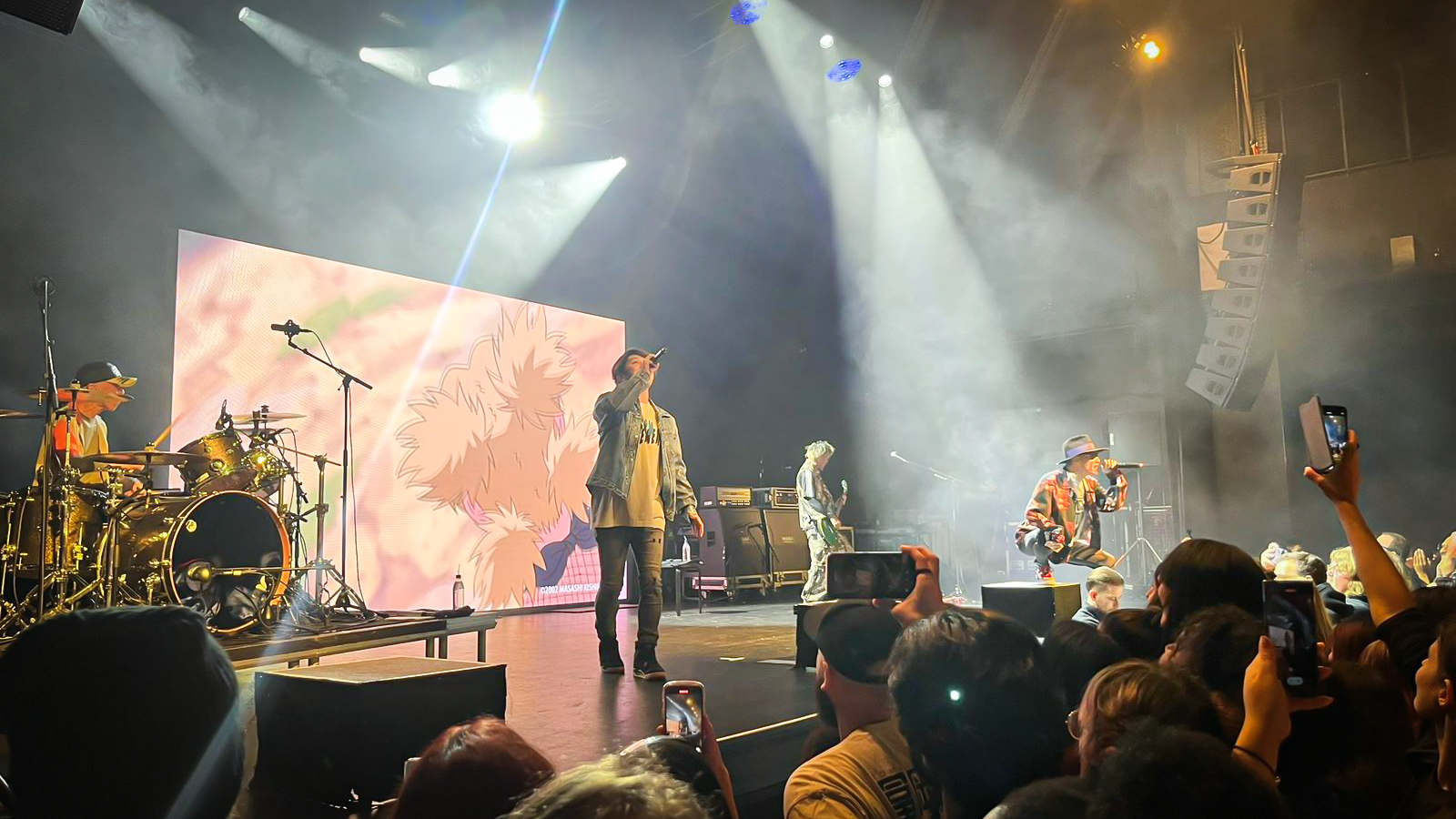 A live performance by the band FLOW, featuring a singer with a microphone and a drummer in the background. The stage is illuminated with dramatic lighting, and an animated character is displayed on a large screen behind the band. The audience is enthusiastically engaged, with some fans recording the performance on their phones.