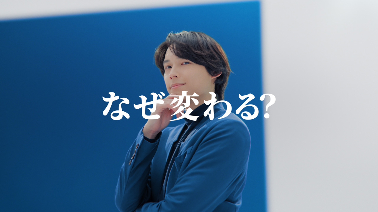 A man in a blue suit poses thoughtfully against a blue background with the text 'なぜ変わる?' (Why change?) displayed prominently.