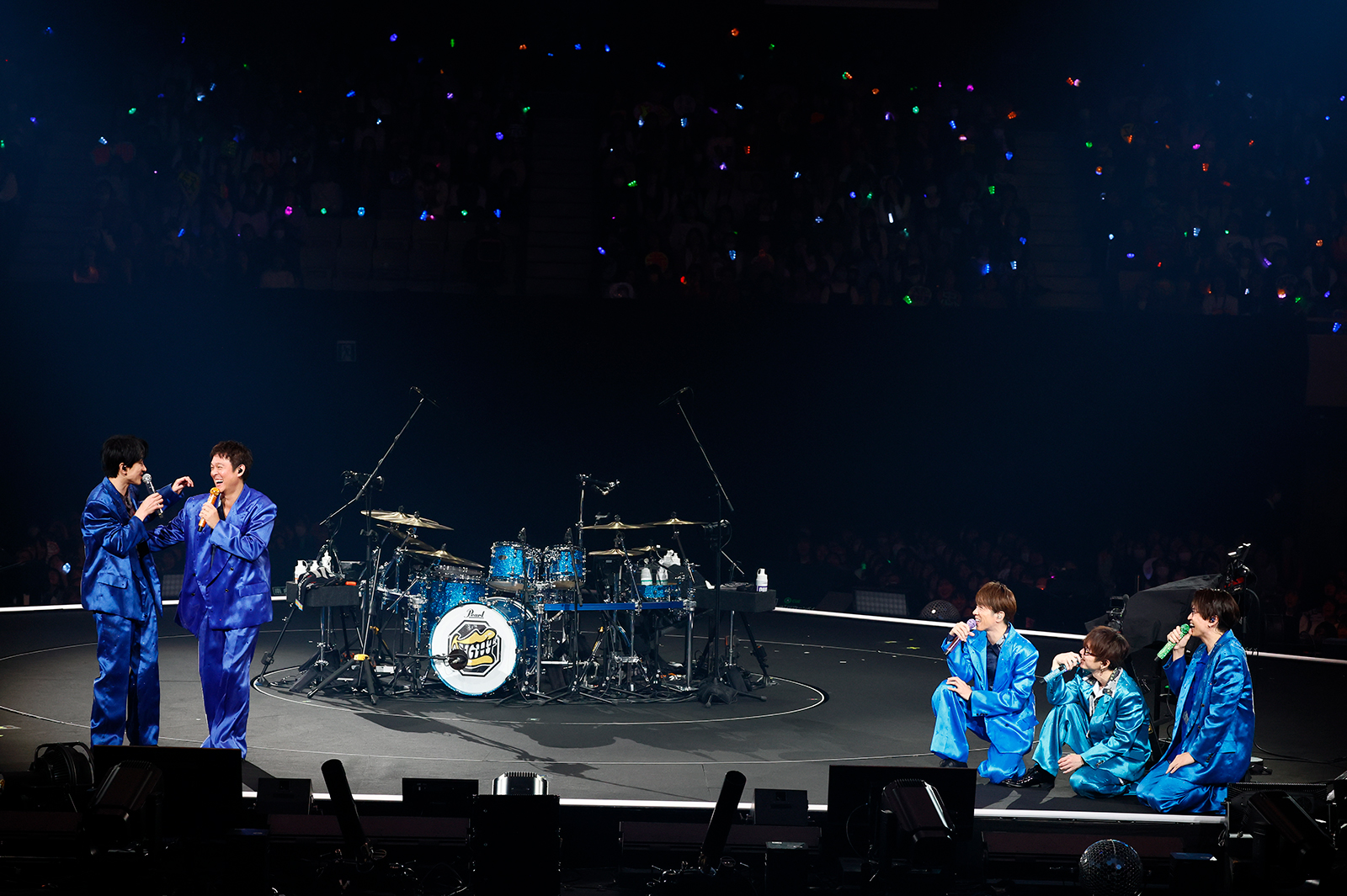 Two members of SUPER EIGHT are speaking into microphones while wearing blue suits, and three other members are crouched nearby, holding microphones. In the background, a drum set is visible, and the audience is filled with fans holding colorful lights.