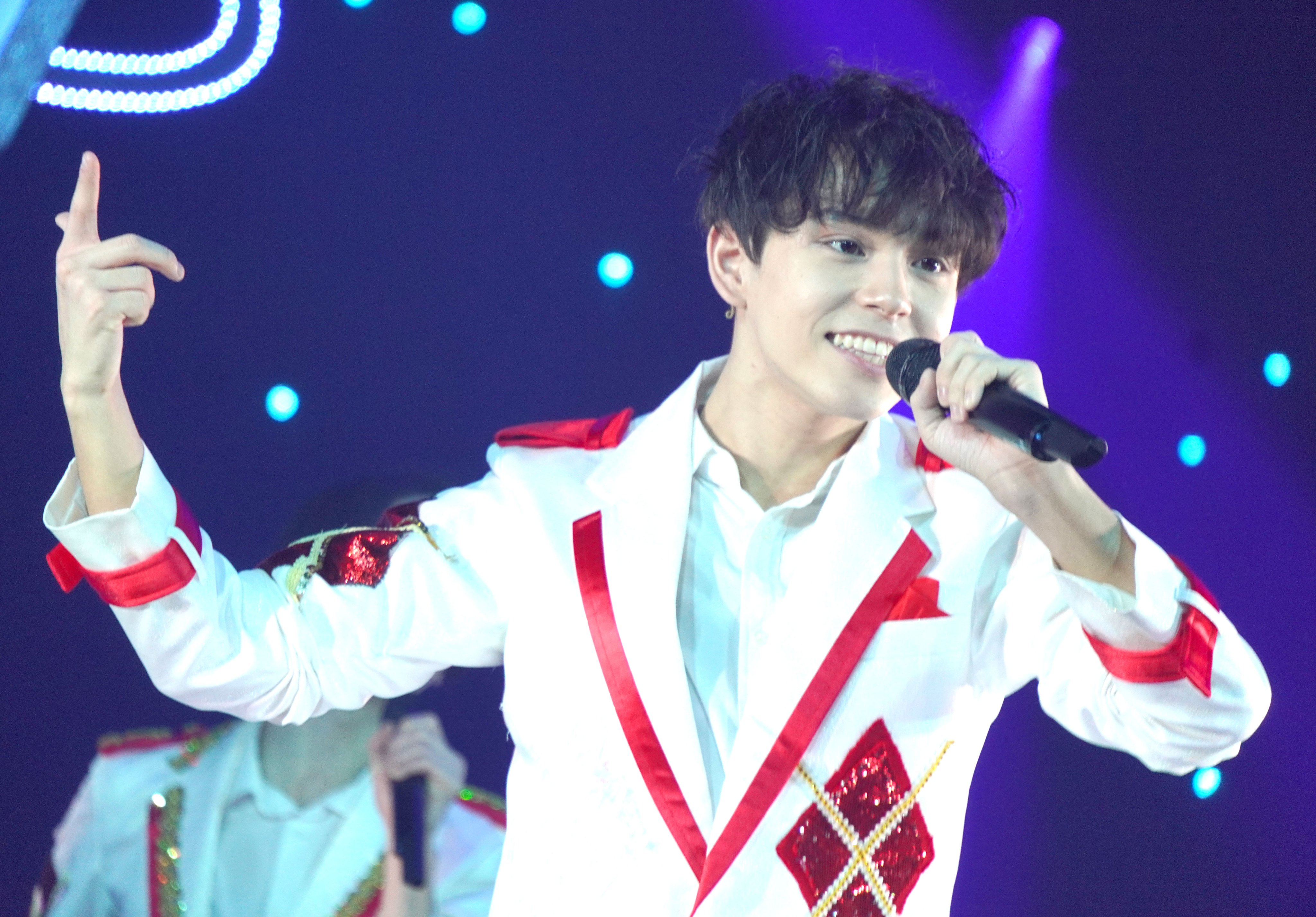 A young performer in a sparkling white and red outfit smiles and gestures while holding a microphone, performing energetically on stage against a colorful, illuminated backdrop.