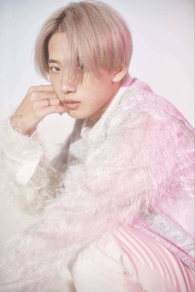 A young person with short, light-colored hair and a thoughtful expression, wearing a stylish, fuzzy white and pink outfit, posed against a softly blurred background.