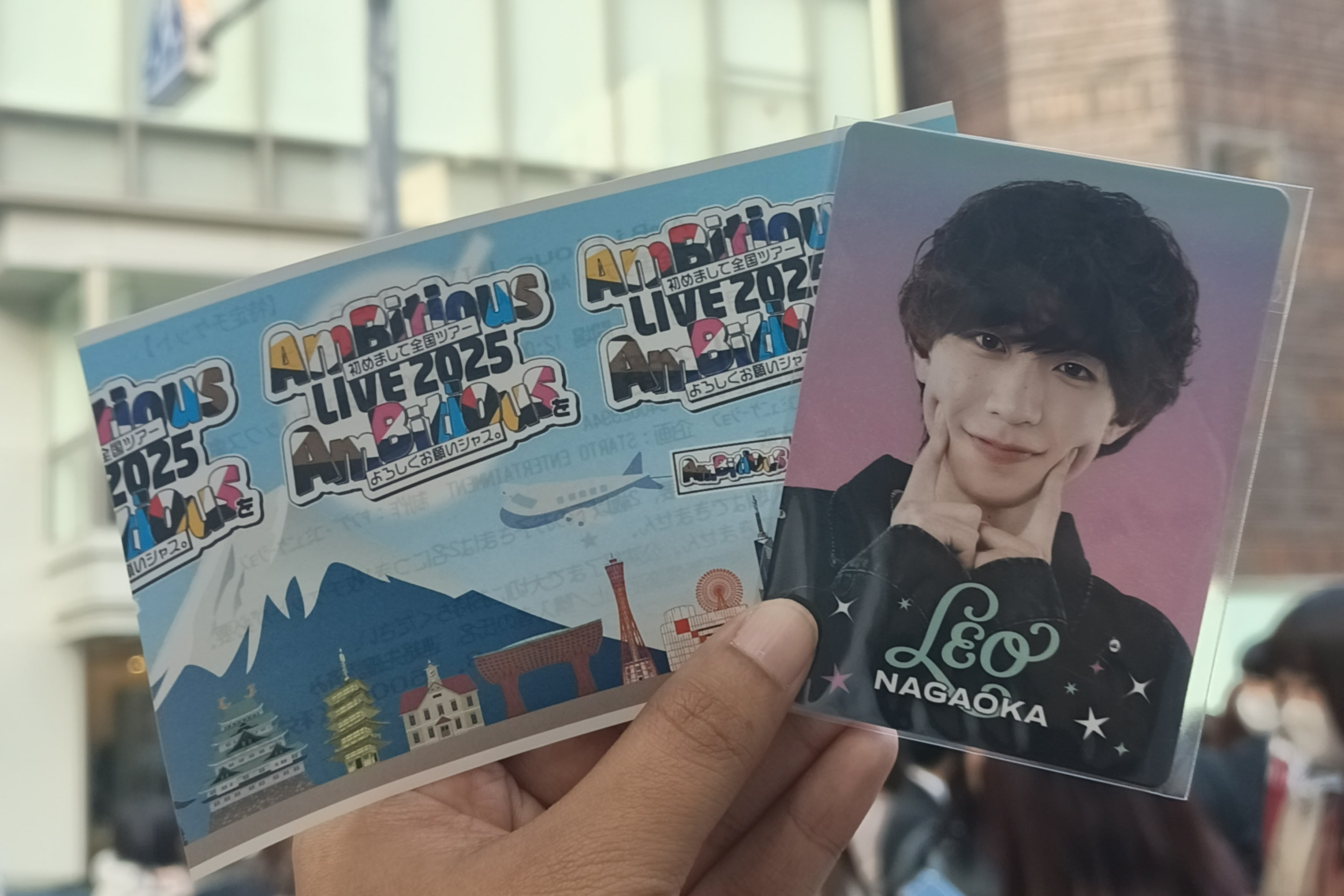 A hand holding two items: a tour pamphlet featuring colorful logos for 'AmBitious LIVE 2025' and a photo card of a member named 'Leo Nagaoka' with a playful expression, against a blurred background.