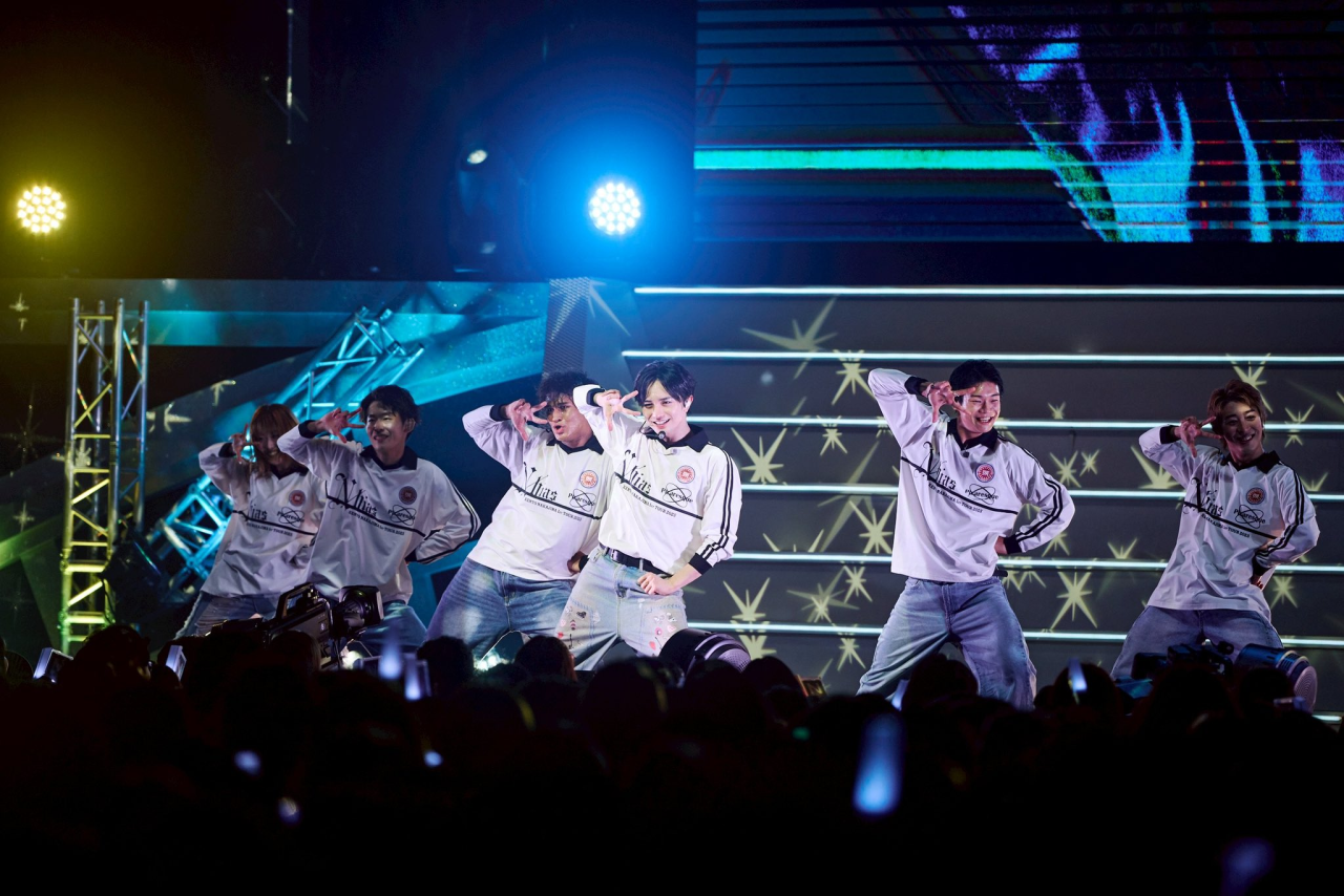 A group of male performers dancing energetically on stage, wearing matching white long-sleeve shirts with logos and denim jeans, under bright stage lights during a live concert.