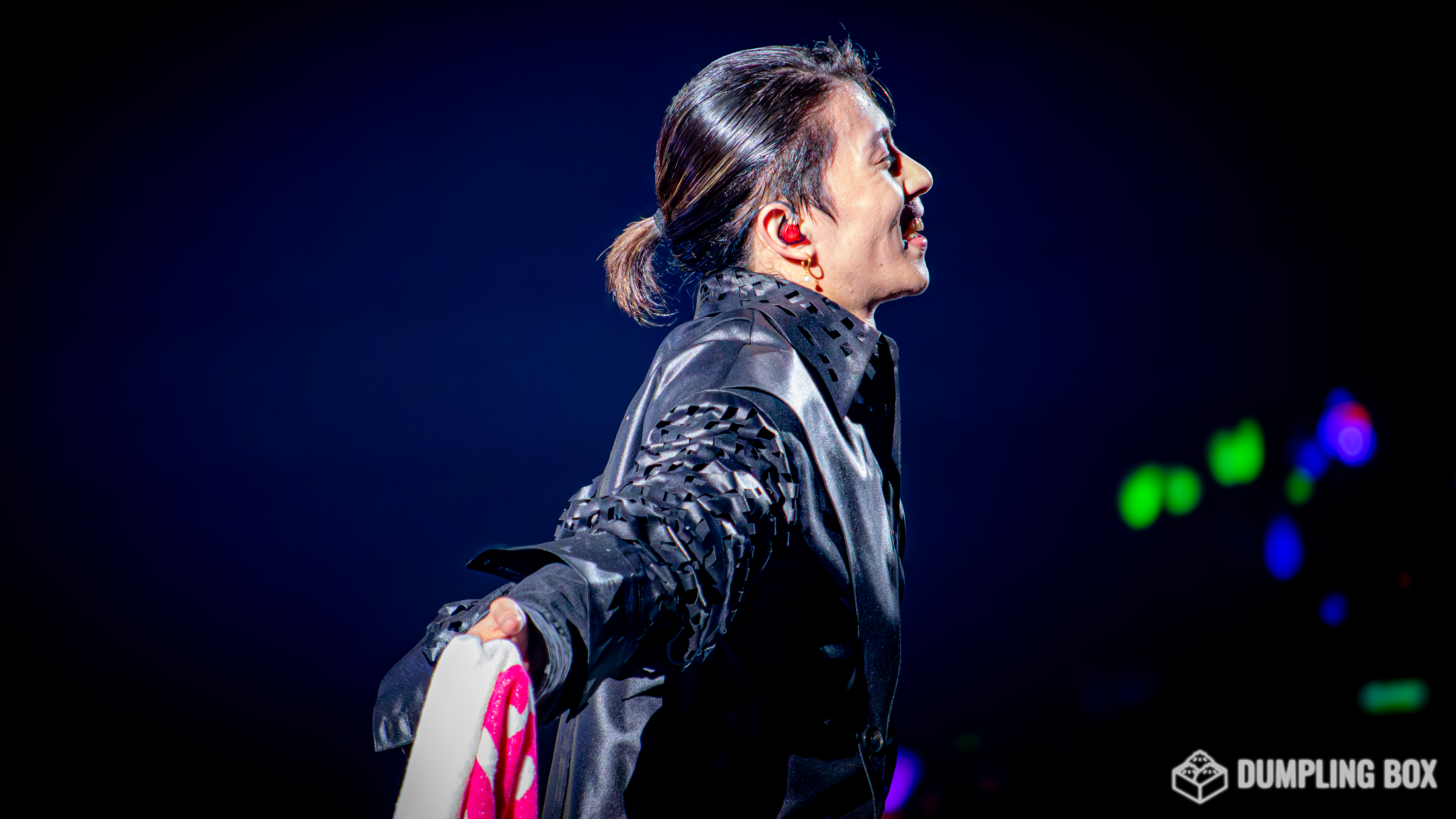 A performer on stage with a joyful expression, dressed in a stylish black outfit with textured sleeves, holding a colorful item. The background features soft lighting effects.