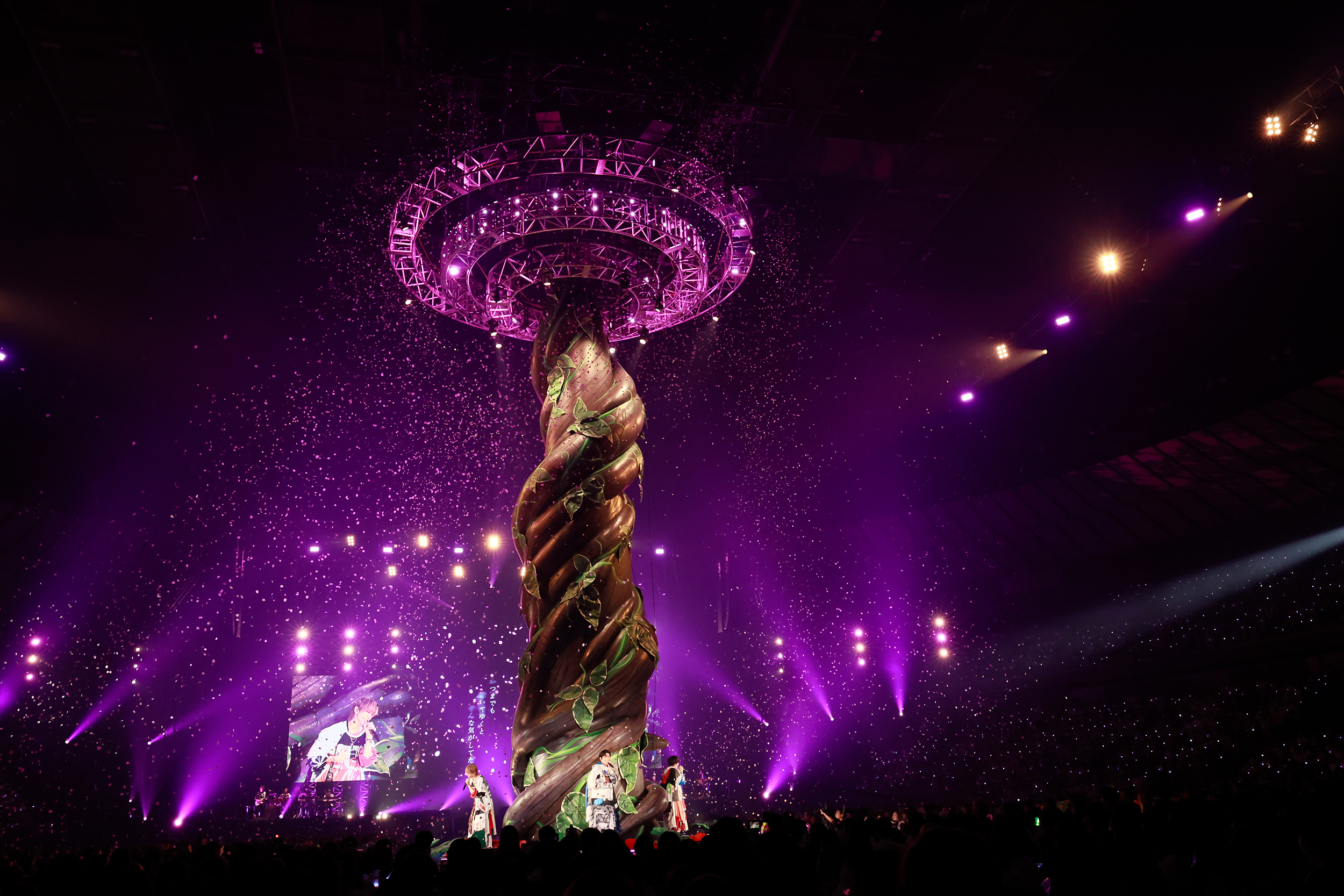 A vibrant stage performance featuring a large, twisted structure resembling a vine with green leaves, illuminated by purple and pink lights, while confetti falls in the air.