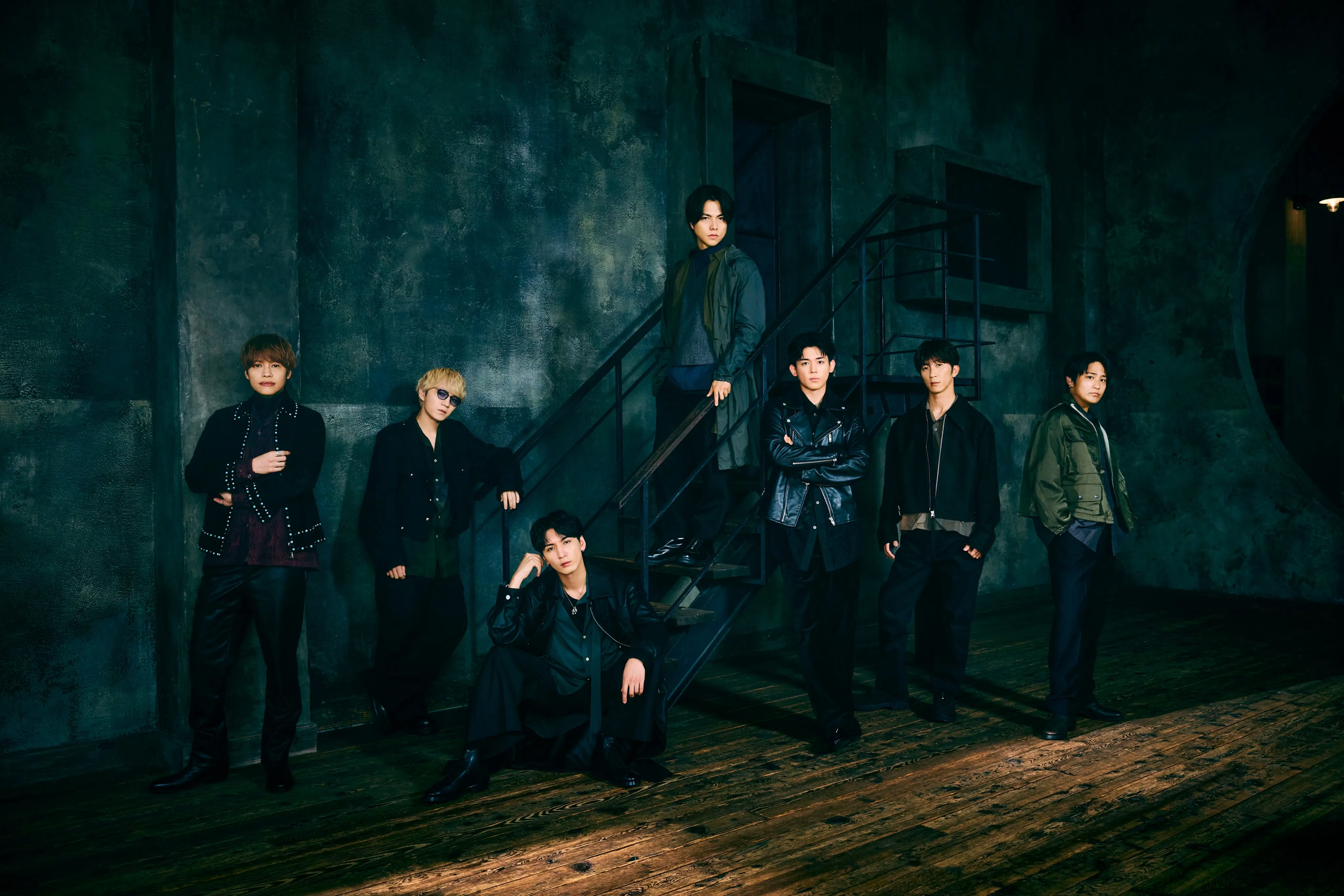 A group of seven individuals posing in a dimly lit industrial setting with a staircase. They are dressed in stylish, dark clothing and are positioned in various stances, some standing and others seated.