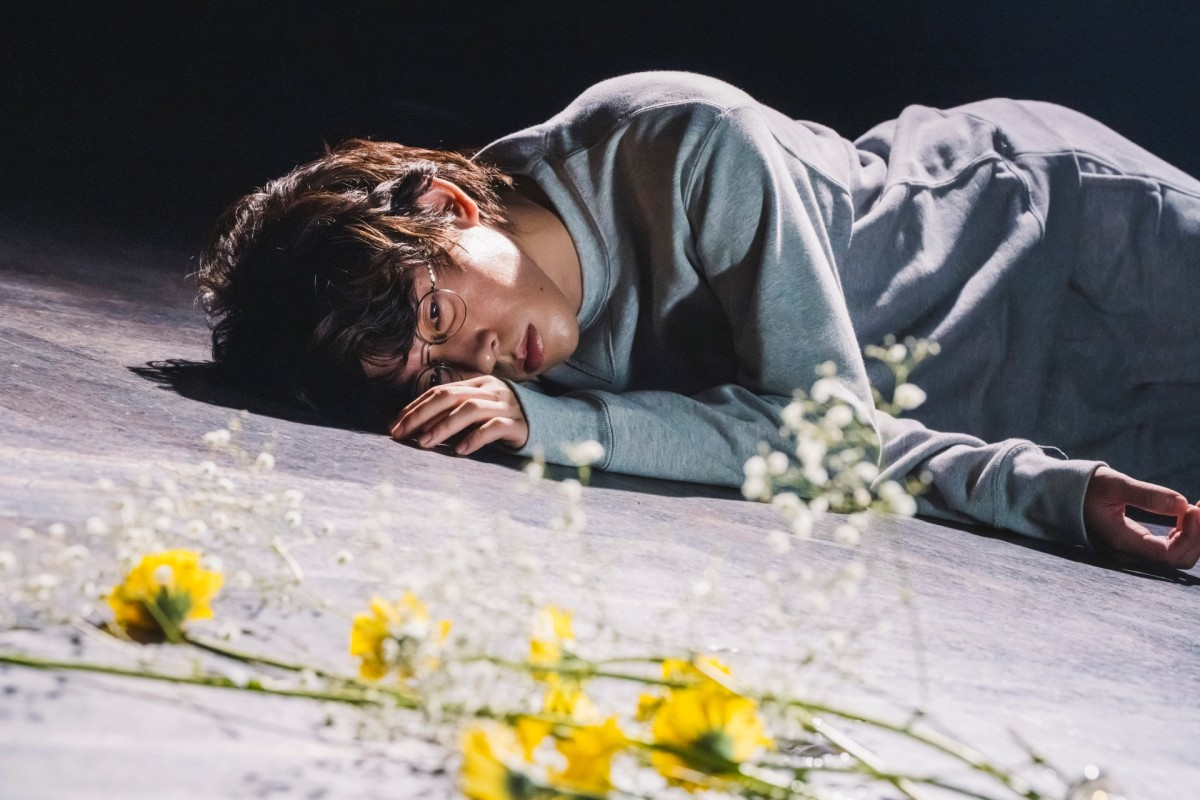 A young person lying on the ground with a pensive expression, wearing a gray sweater and glasses, surrounded by yellow flowers and baby's breath.