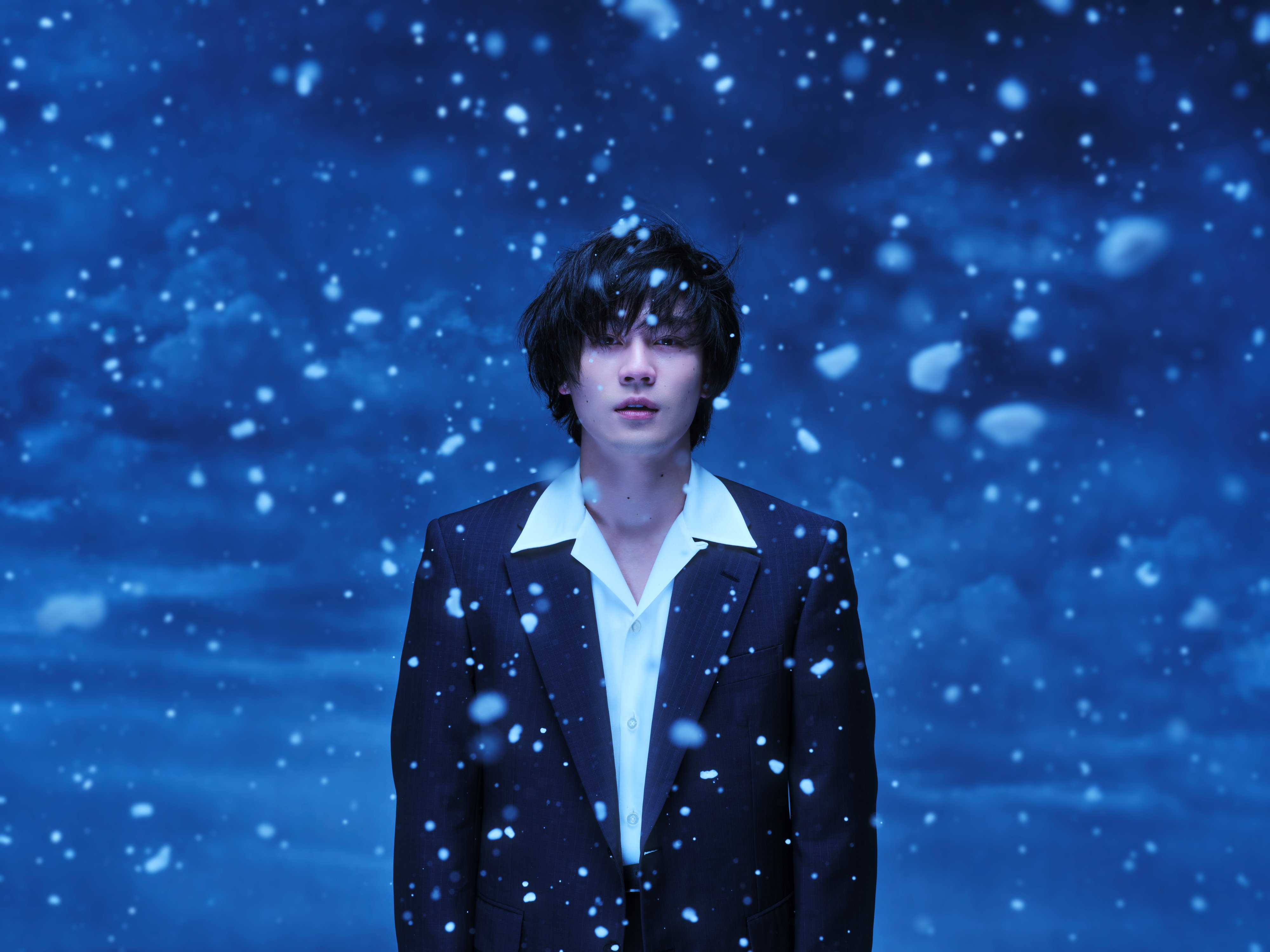 A person in a dark suit stands amid falling snow against a blue background, with a somber expression.