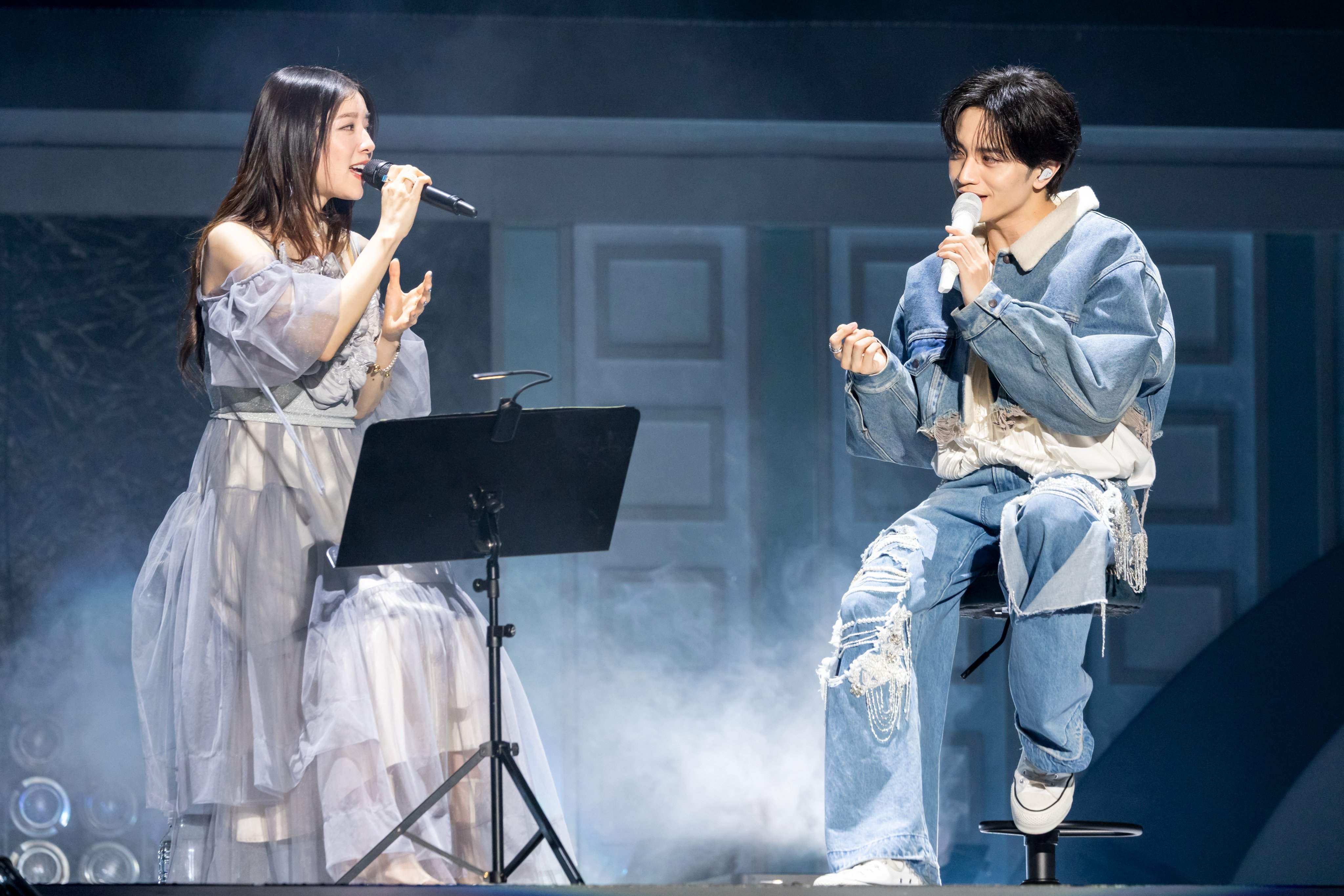 A female vocalist in a flowing gray dress sings into a microphone while a male vocalist in denim attire sits on a stool, engaged in the performance. The stage is filled with soft lighting and haze, creating an atmospheric setting.
