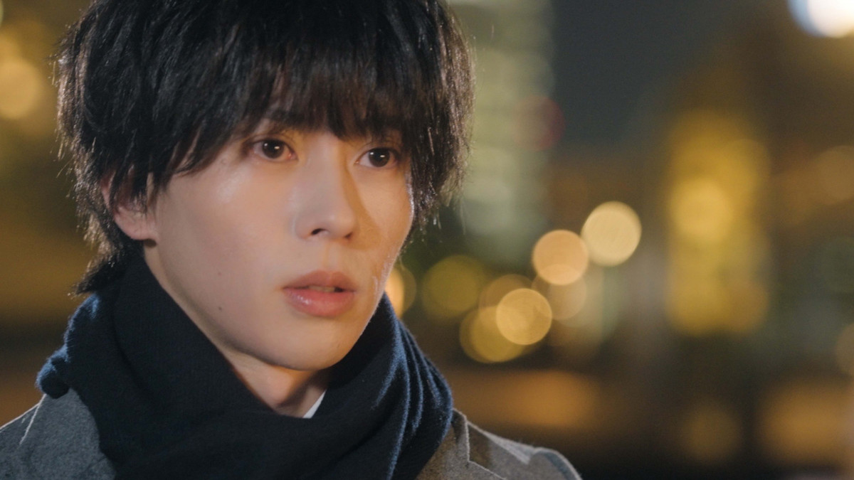 Close-up of a young man with dark, tousled hair and a serious expression, wearing a scarf, against a blurred background of city lights at night.