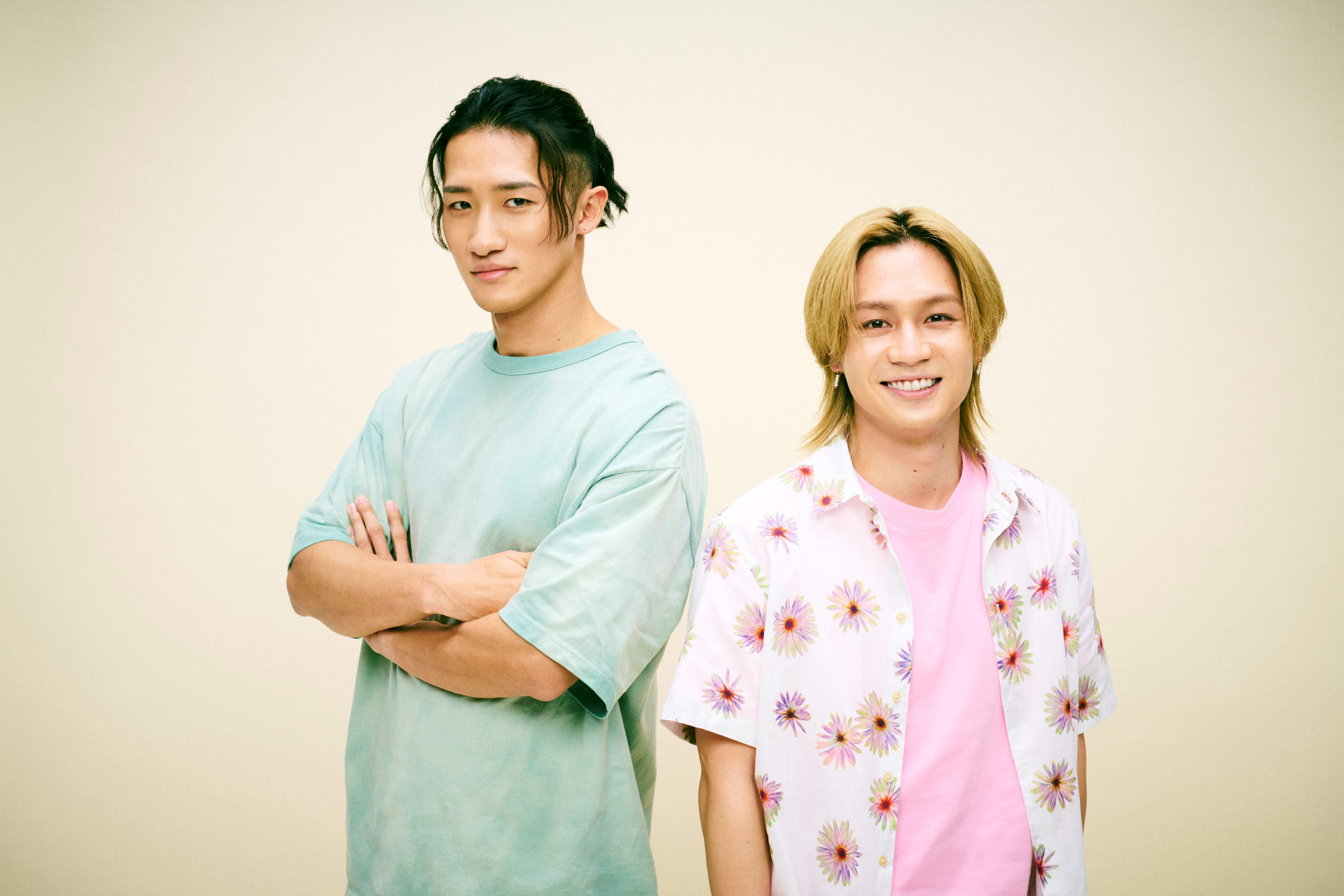 Two young men standing against a light yellow background. One is wearing a mint green oversized t-shirt with arms crossed and has long dark hair. The other is smiling and wearing a pink shirt under a floral-patterned short-sleeve shirt.