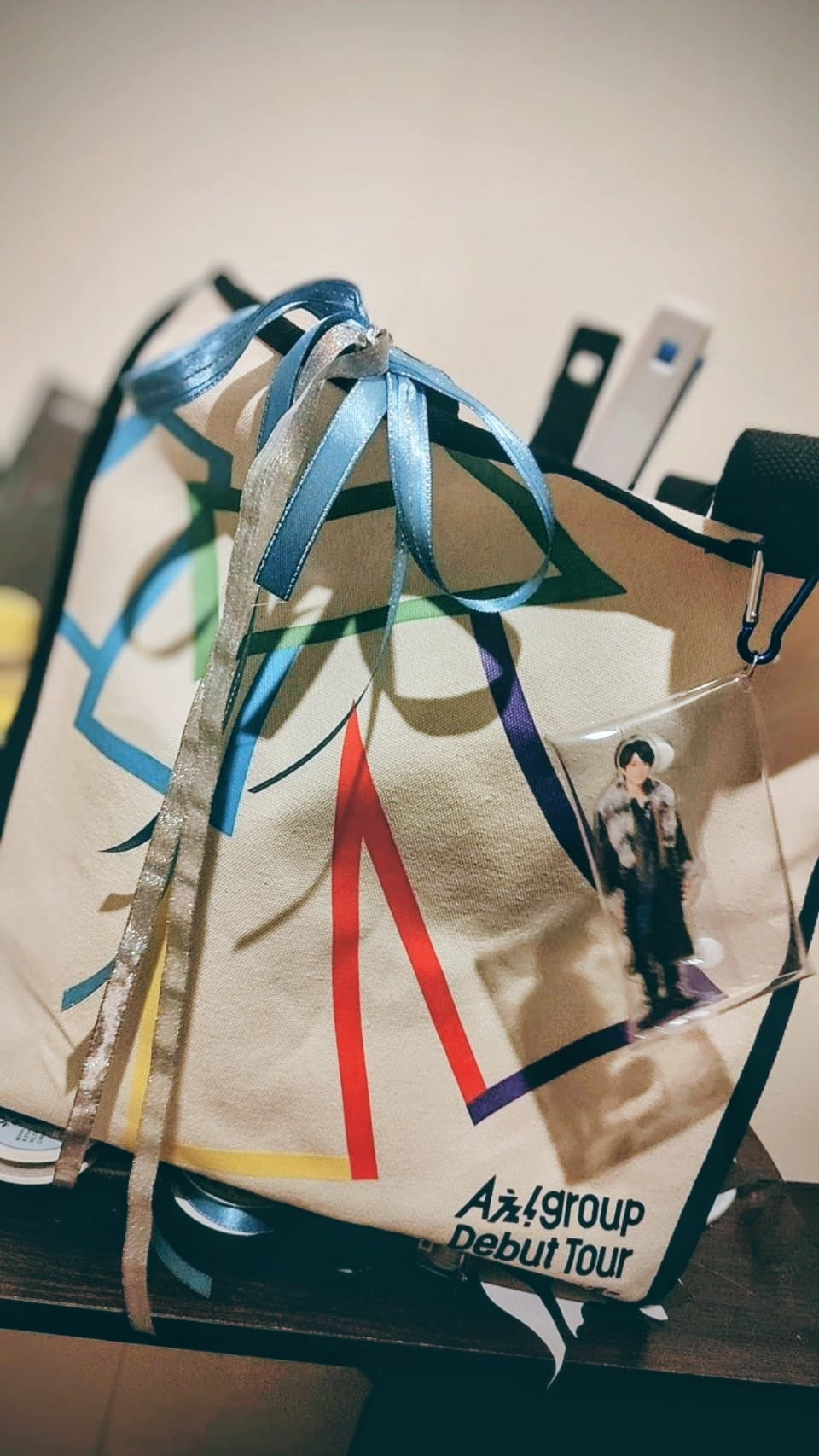 A tote bag featuring colorful geometric designs and ribbons, with a clear charm depicting a character attached to it. The bag has text that reads 'A'z group Debut Tour'.