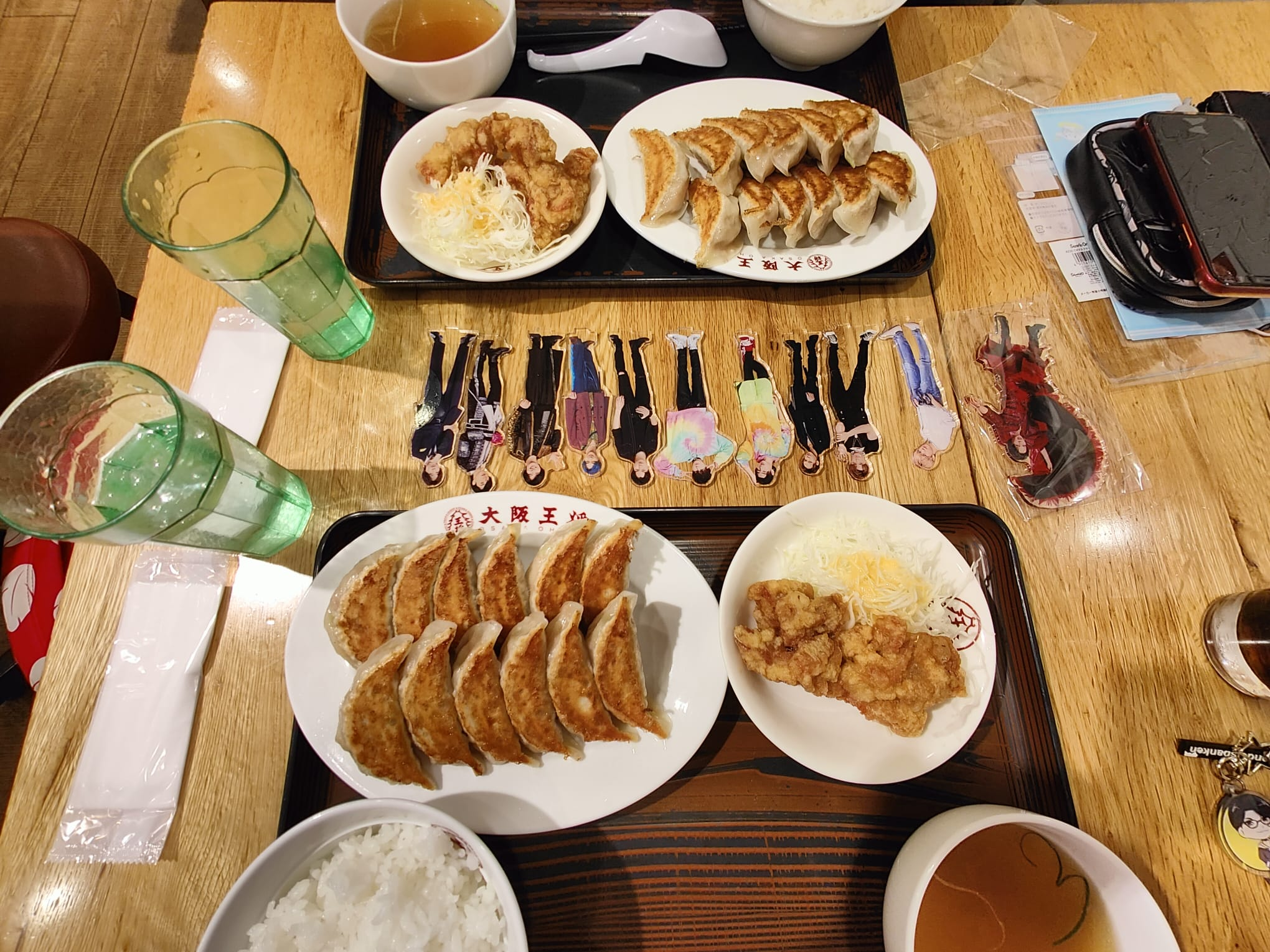 A top-down view of a dining table with two plates of gyoza, a bowl of rice, and a dish of fried chicken, accompanied by two glasses of green beverage and decorative paper cut-outs of characters.