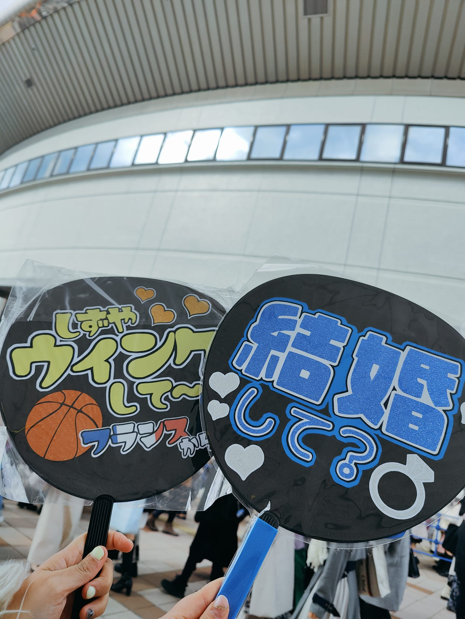 Two hand-held fans with playful messages in Japanese, one featuring a basketball theme and the other asking about marriage, displayed against a backdrop of a modern building.