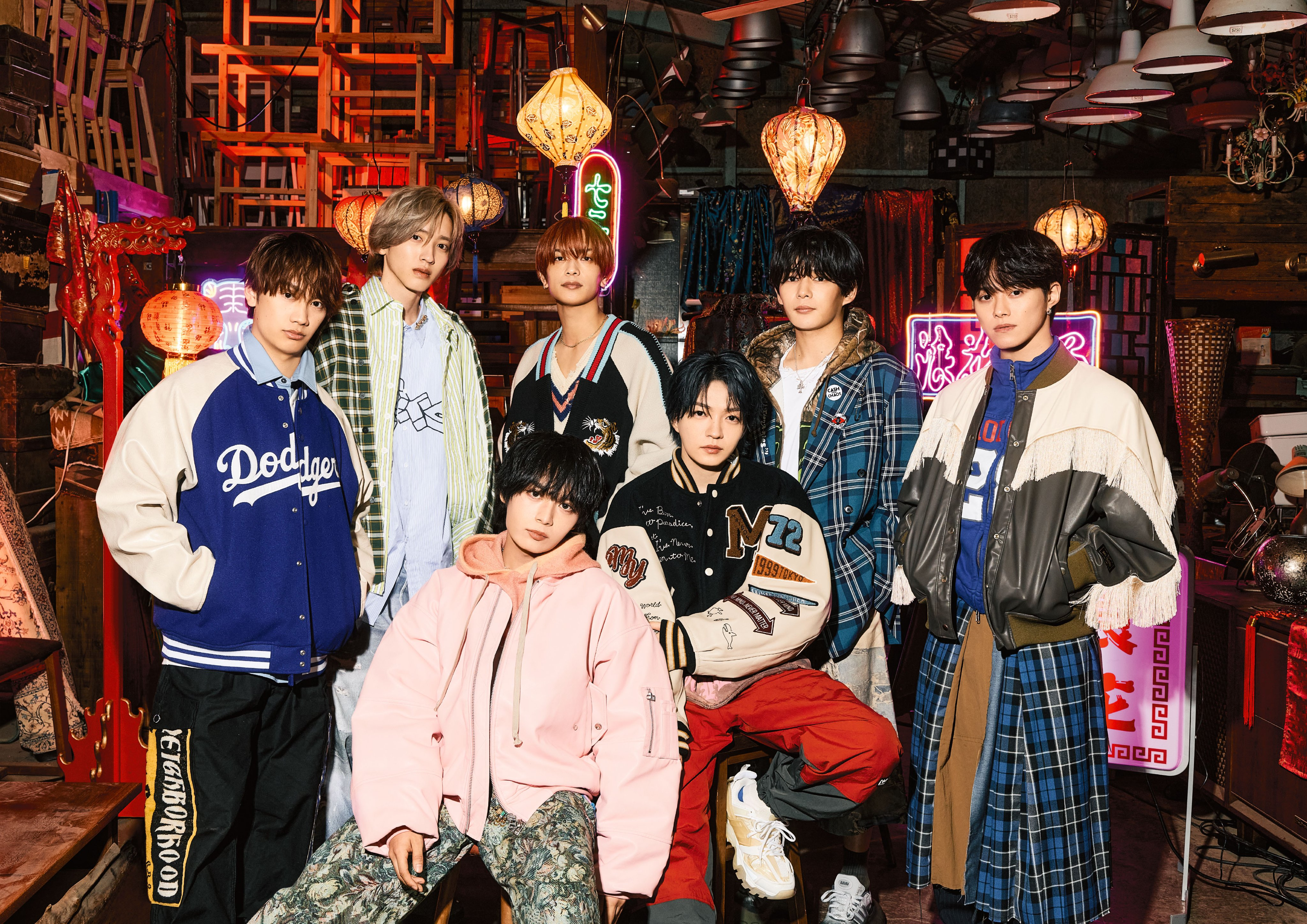 A group photo of seven members of Naniwa Danshi posing together in a colorful, eclectic setting with lanterns and neon lights in the background. They are dressed in a mix of stylish and casual outfits.