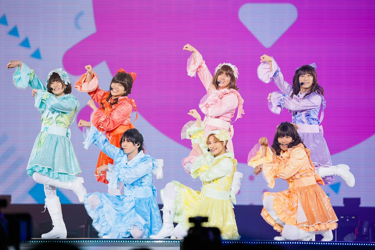 A group of performers dressed in colorful, sparkling outfits, posing energetically on stage during a concert with a vibrant background.