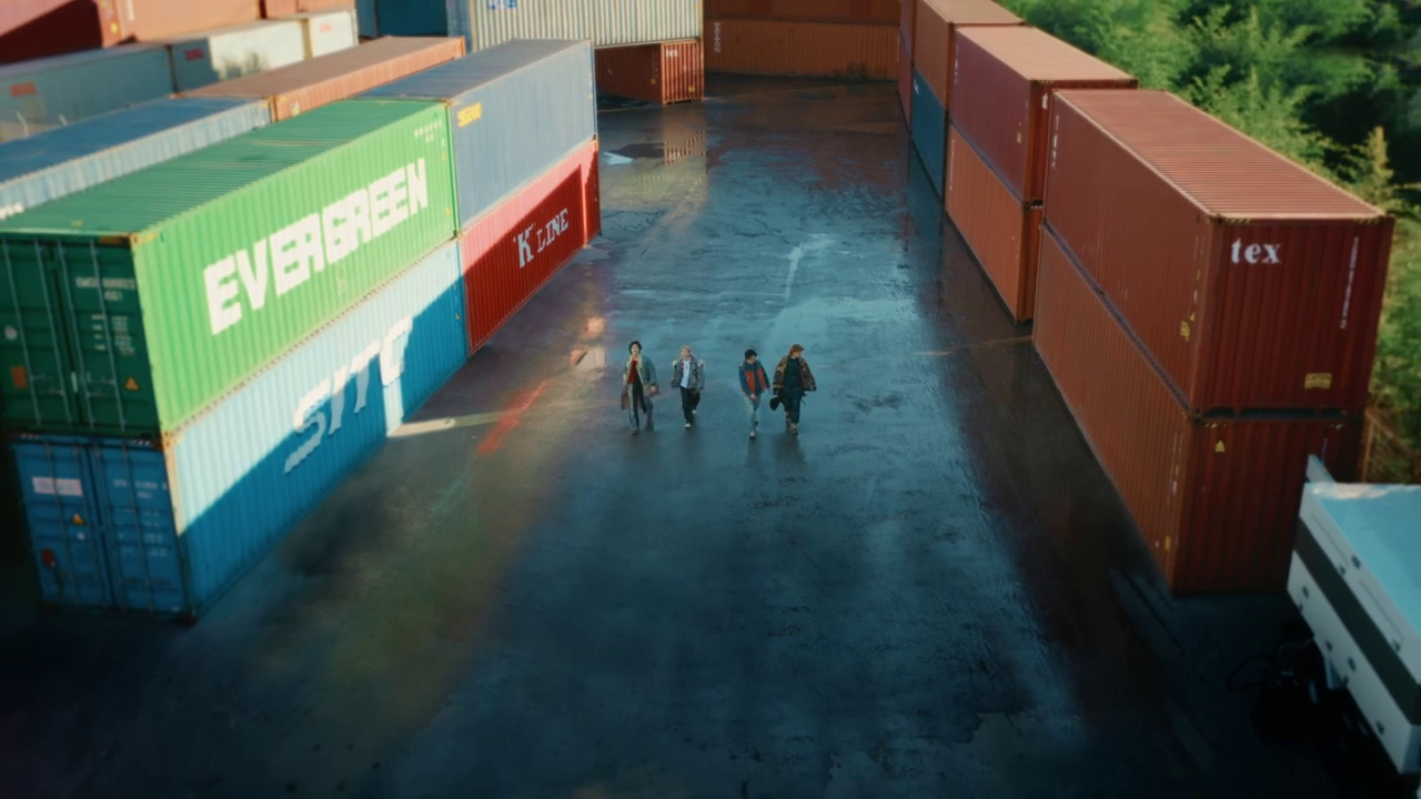 Four individuals walking on a wet surface between colorful shipping containers in a yard.