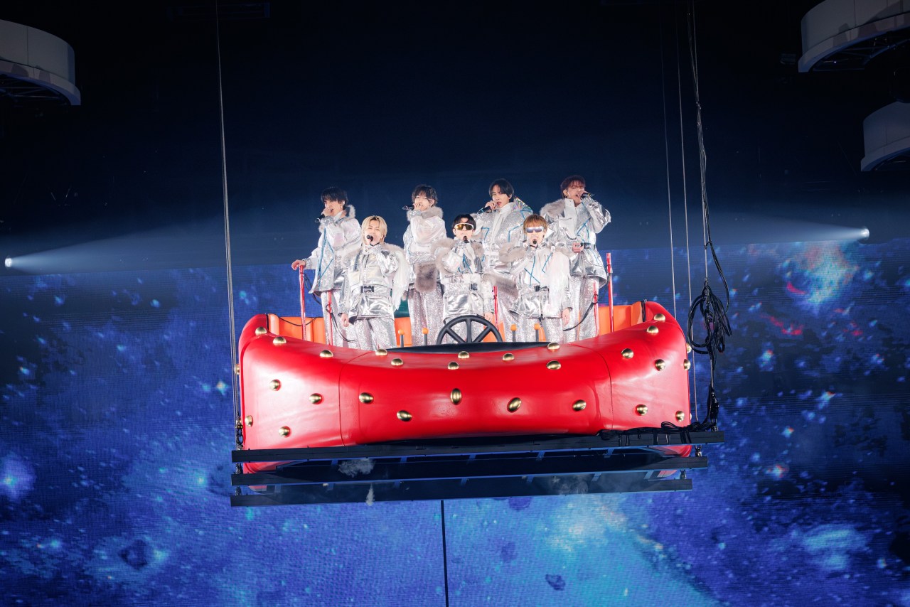 A group of performers dressed in shiny silver outfits stands on a large red platform resembling a spacecraft, suspended above a cosmic background with blue and purple hues.