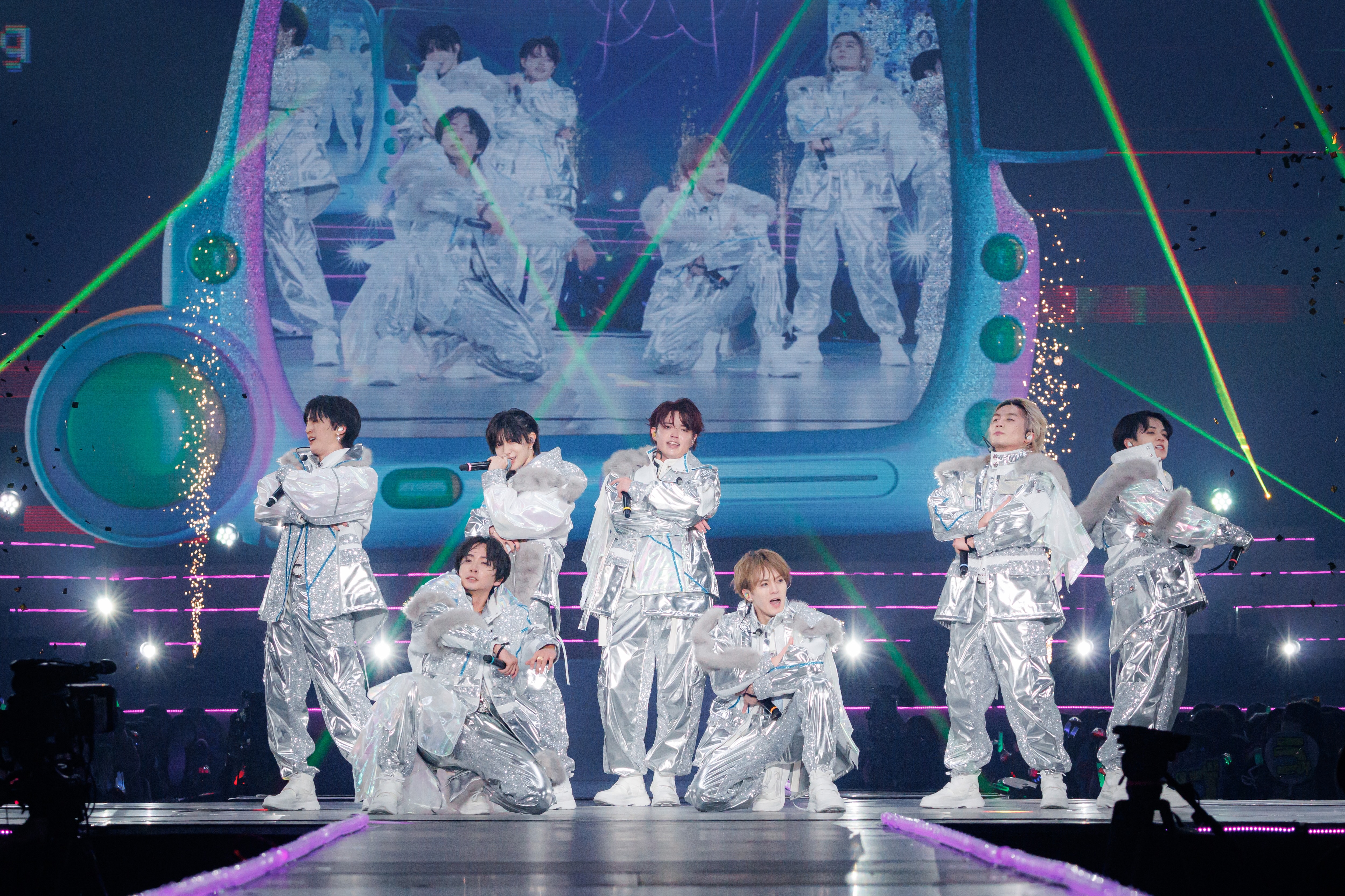 A group of performers in shiny silver outfits performing on stage, with colorful lights and a large screen displaying their image in the background. Confetti and sparkles add to the vibrant atmosphere.
