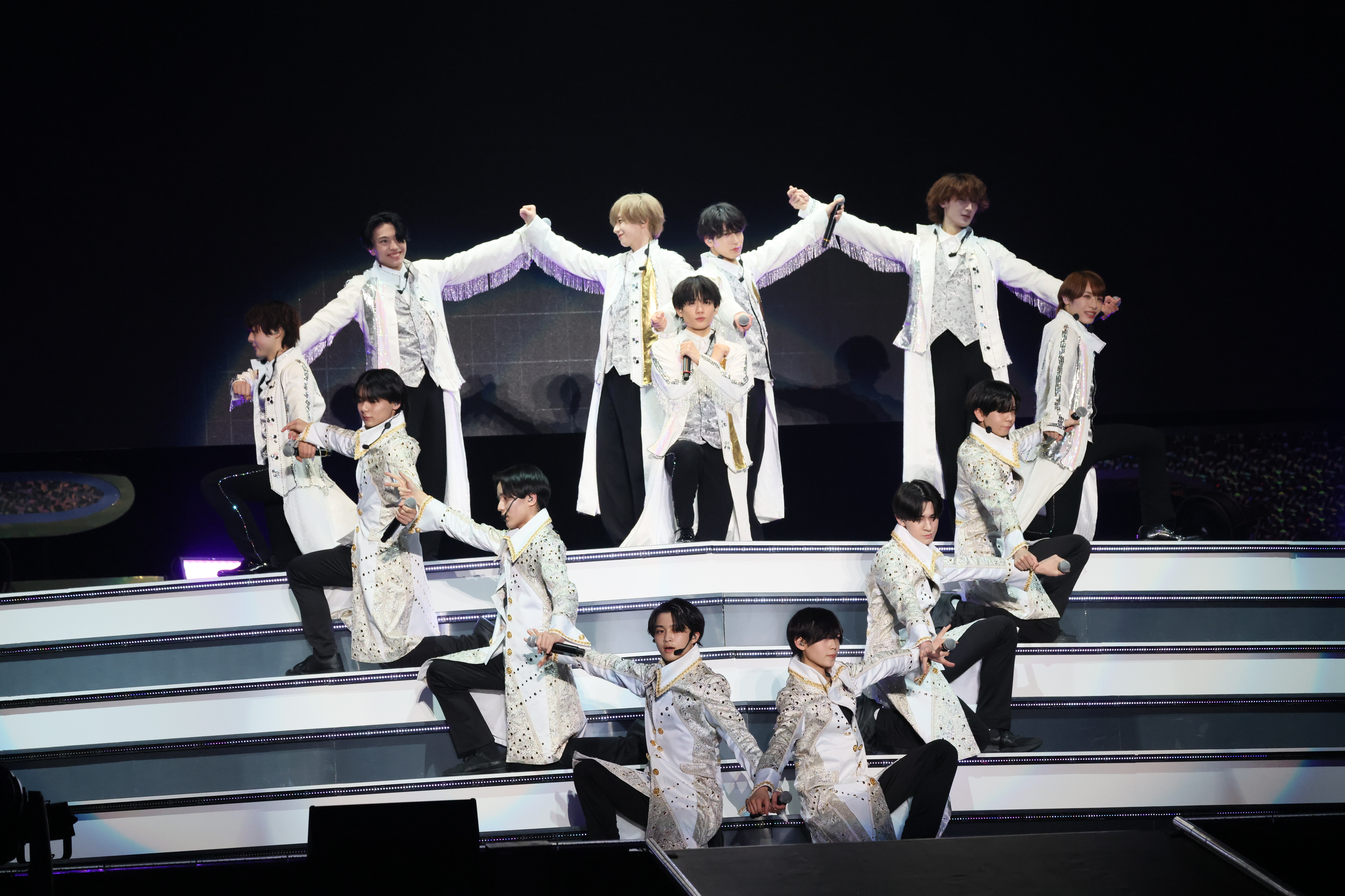 A group of performers dressed in matching white and silver outfits striking dynamic poses on stage, with two performers standing prominently at the back and others seated or crouched in front.