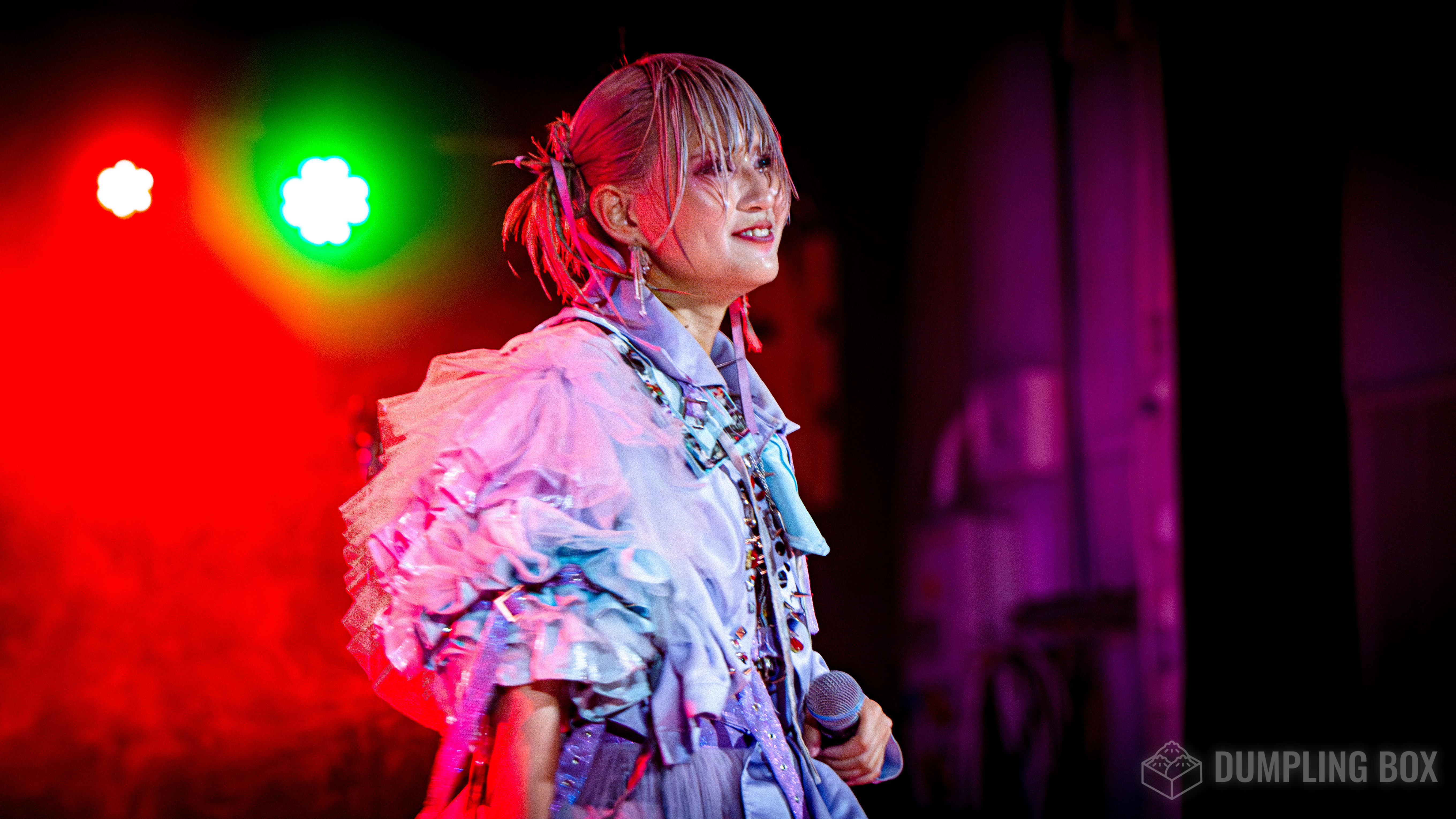 A performer smiling on stage, wearing a colorful, ruffled outfit, holding a microphone, with vibrant stage lights in the background.