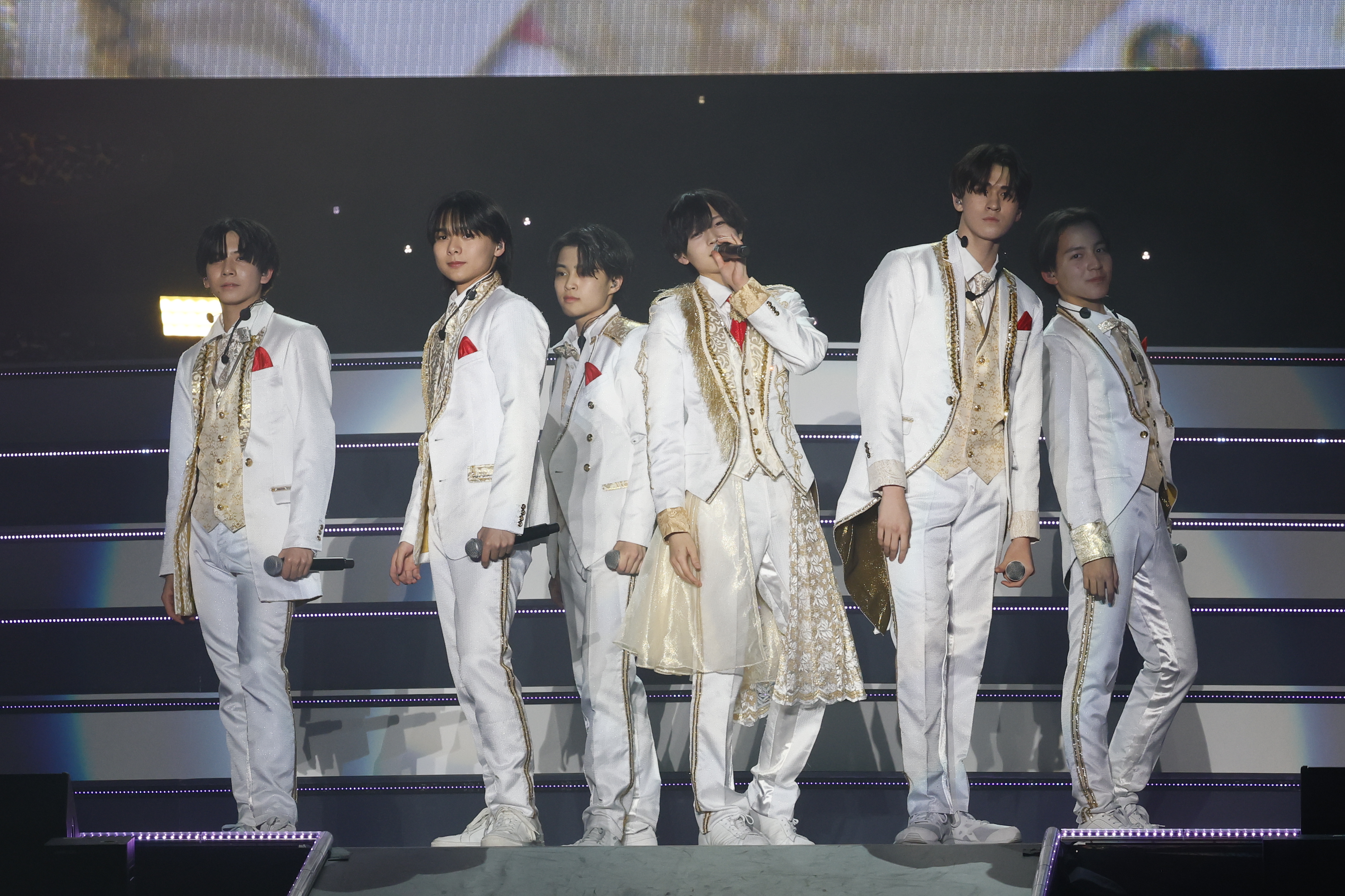 A group of six performers dressed in white and gold costumes with red accents, posing on stage during a concert.
