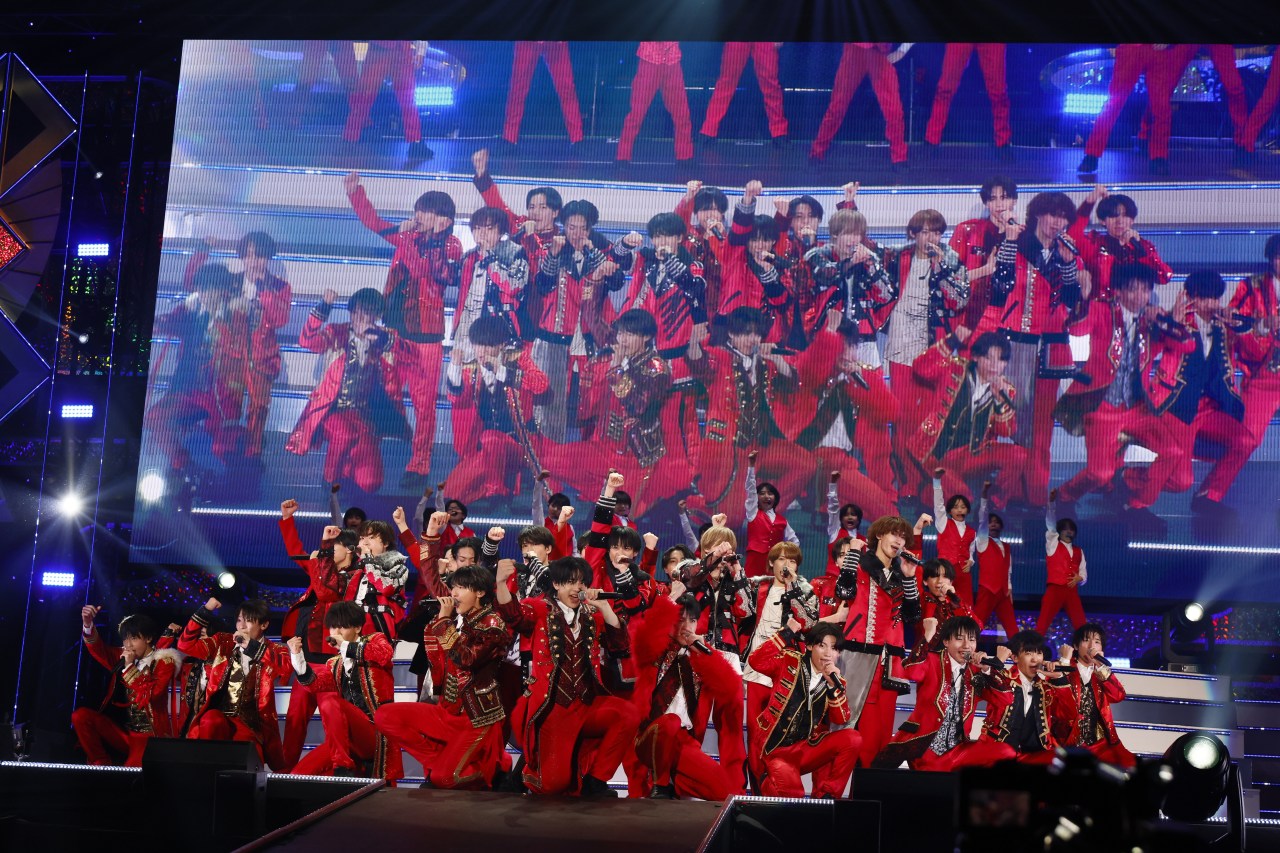A group of performers dressed in red costumes poses dynamically on stage, with a large screen displaying their image behind them, showcasing a vibrant concert atmosphere.