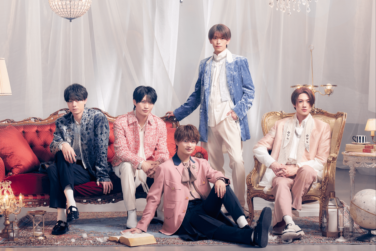 A group of five elegantly dressed individuals posing together in a stylish interior setting. They are seated on a red velvet couch, with decorative elements like a chandelier, a lamp, and a globe in the background. Each person wears unique, fashionable clothing with pastel colors and textures.