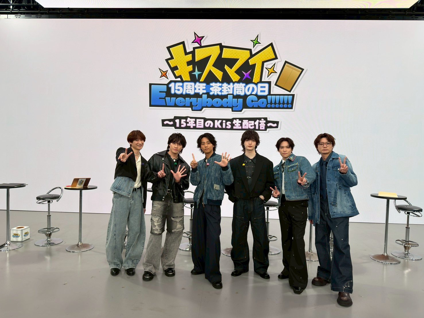 A group of six individuals stands together in front of a large screen displaying the event title in Japanese characters, celebrating the 15th anniversary of a band. They are dressed in denim outfits and making peace signs.