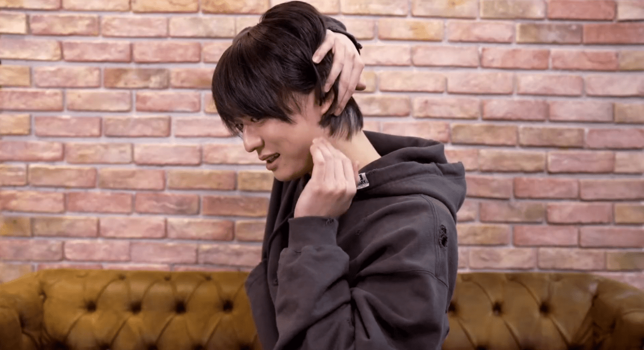 A young person with dark hair is sitting on a brown tufted couch, touching their neck while looking to the side. The background features a brick wall.