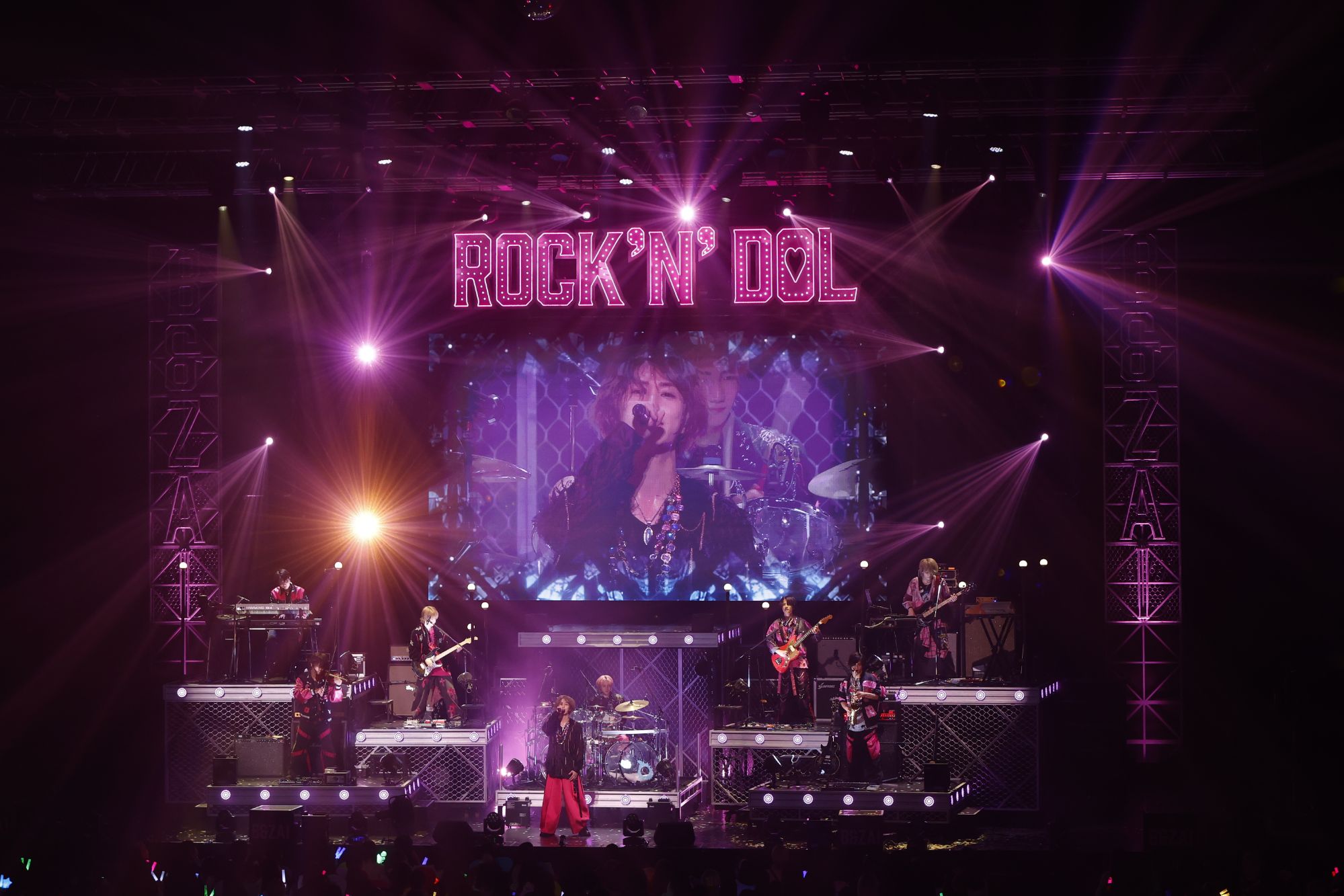 A live concert performance featuring a band on stage with colorful lighting and a large video screen behind them. The band members are dressed in stylish outfits, with one vocalist taking center stage.