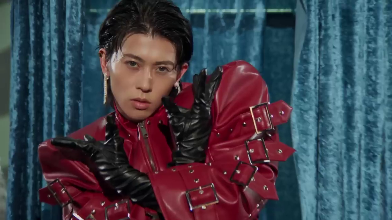 A person poses dramatically in a red leather jacket with buckles and black gloves, showcasing a bold fashion style against a blue backdrop.