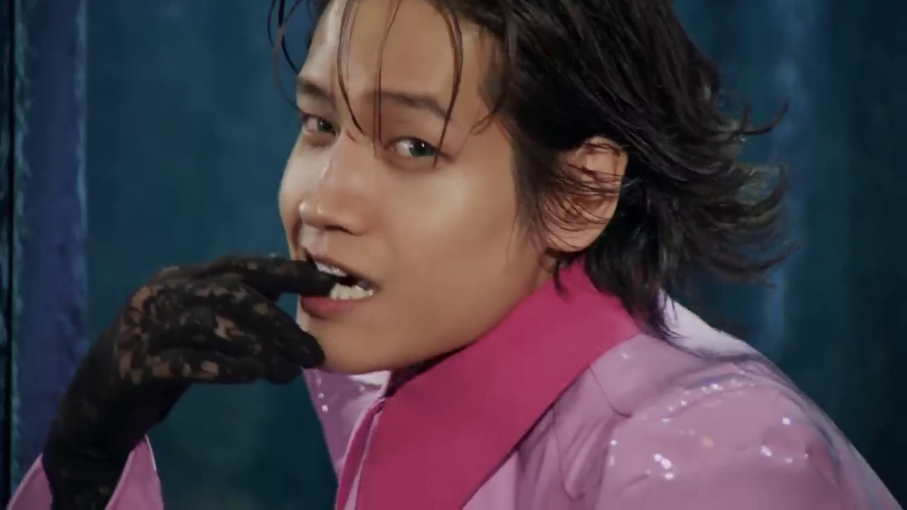 A person with tousled hair, wearing a pink outfit and black lace gloves, bites their finger while looking playfully at the camera.