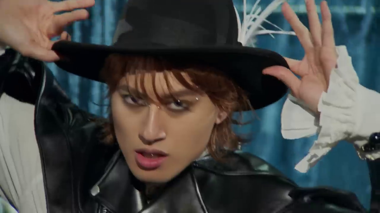 A person with tousled hair wearing a black hat and leather jacket, posing confidently against a blue background.