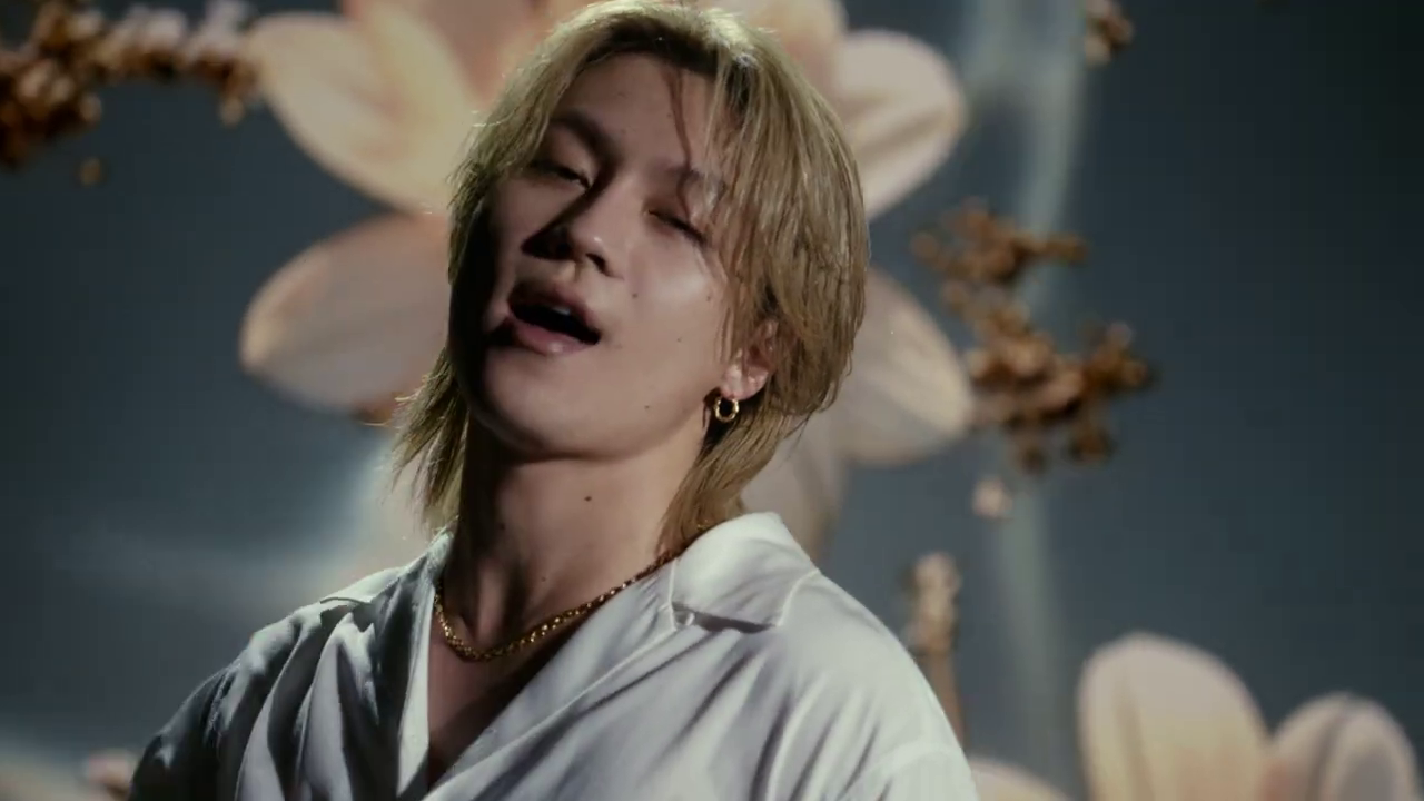A young male singer with long, light-colored hair and wearing a white shirt is singing passionately against a backdrop of soft floral patterns.