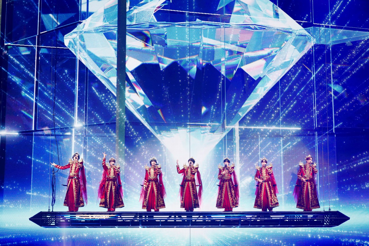 A performance stage featuring a group of eight performers dressed in ornate red and gold costumes, standing on a platform. Behind them, a large diamond-shaped structure is illuminated with sparkling lights, creating a vibrant visual display.
