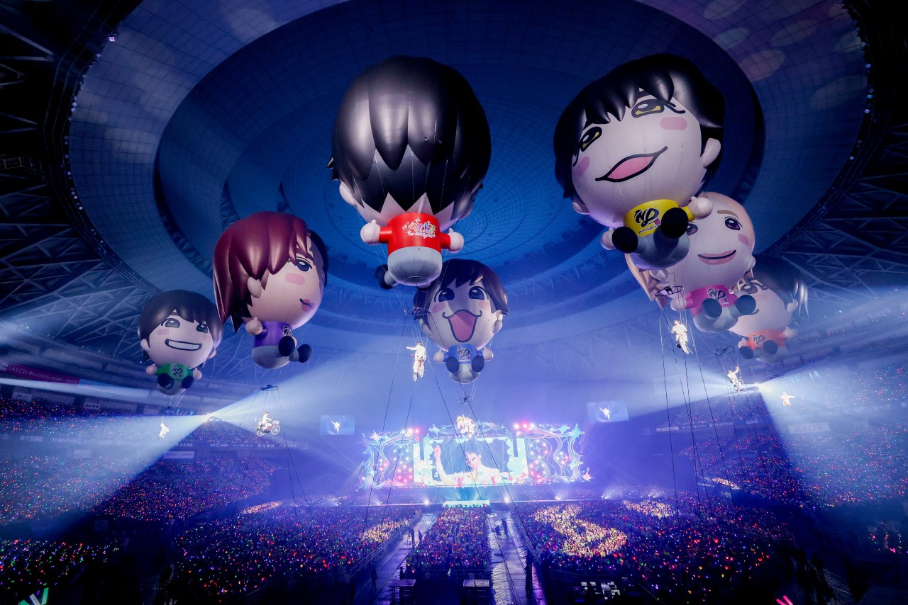 A colorful concert scene featuring large inflatable character balloons floating above an enthusiastic crowd in a stadium, with vibrant lights illuminating the venue.