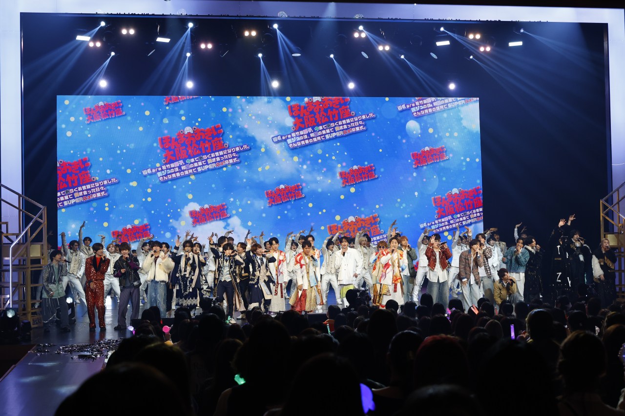A colorful stage performance featuring a large group of performers in elaborate costumes, standing together and interacting with an enthusiastic audience. The backdrop features vibrant graphics and text, enhancing the festive atmosphere.